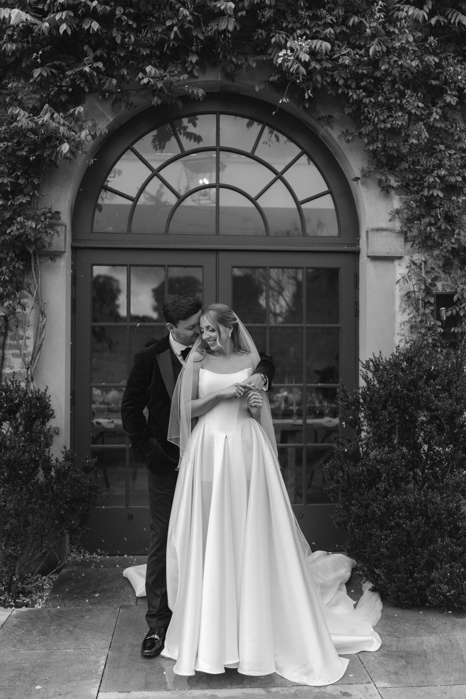 Black and white photo of a bride and groom embracing before an arched window covered in ivy at their wedding venue.