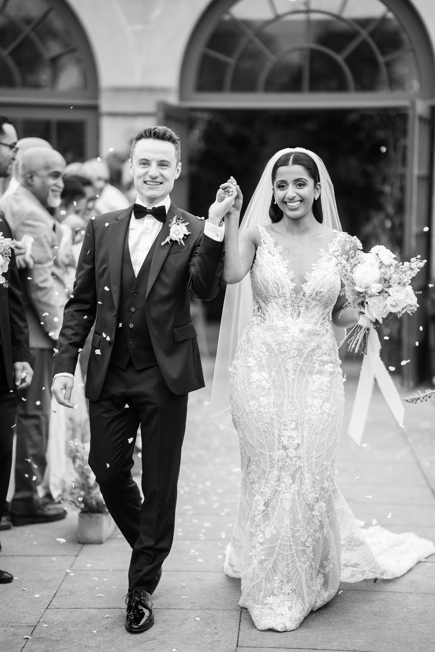 Black and white photo of a smiling bride and groom walking out of a venue with confetti falling around them.