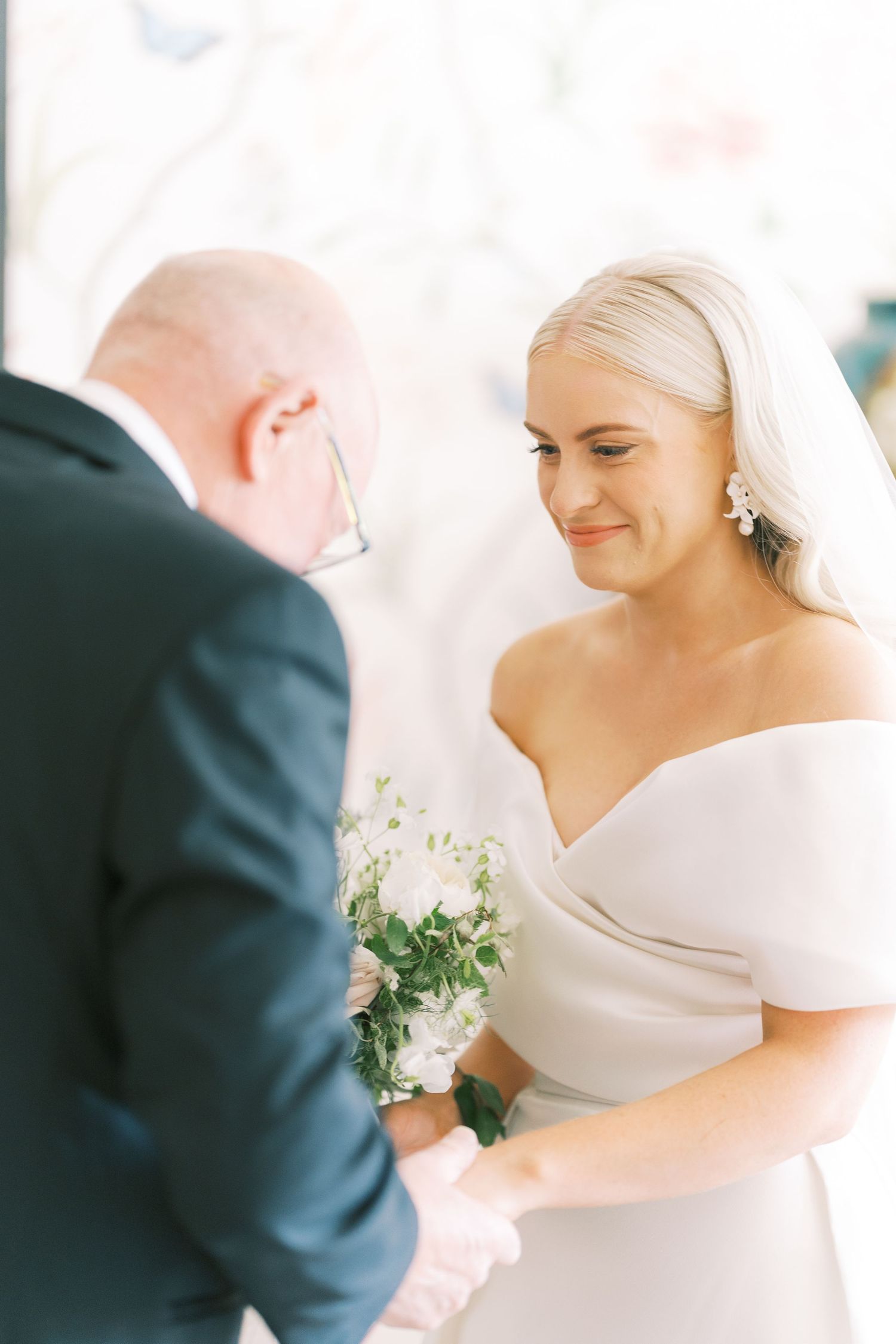 Emotional bride in off-shoulder gown smiling during wedding ceremony while holding hands with officiant.