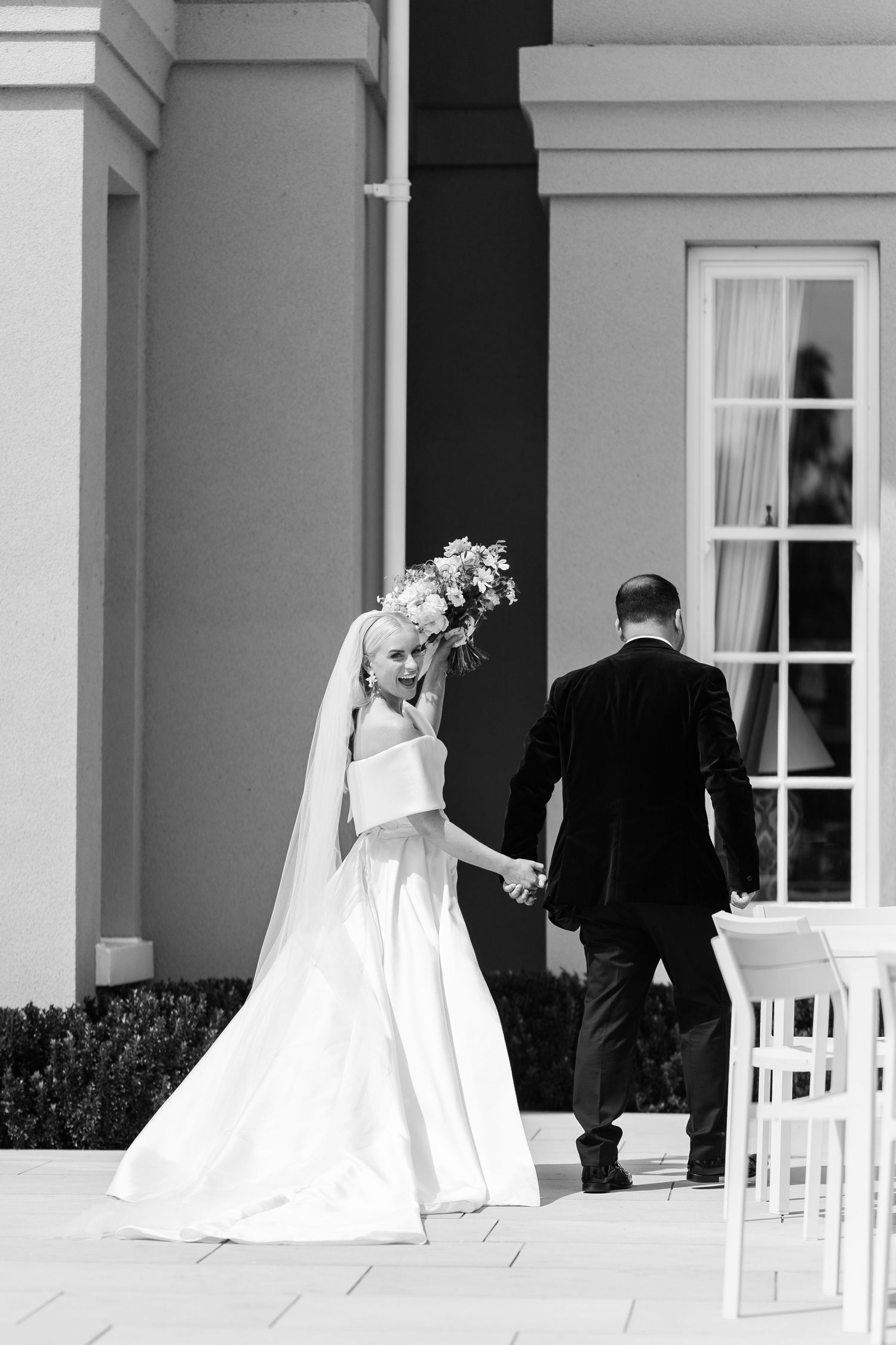Bride in off-shoulder gown holds bouquet while walking hand-in-hand with groom toward elegant venue.