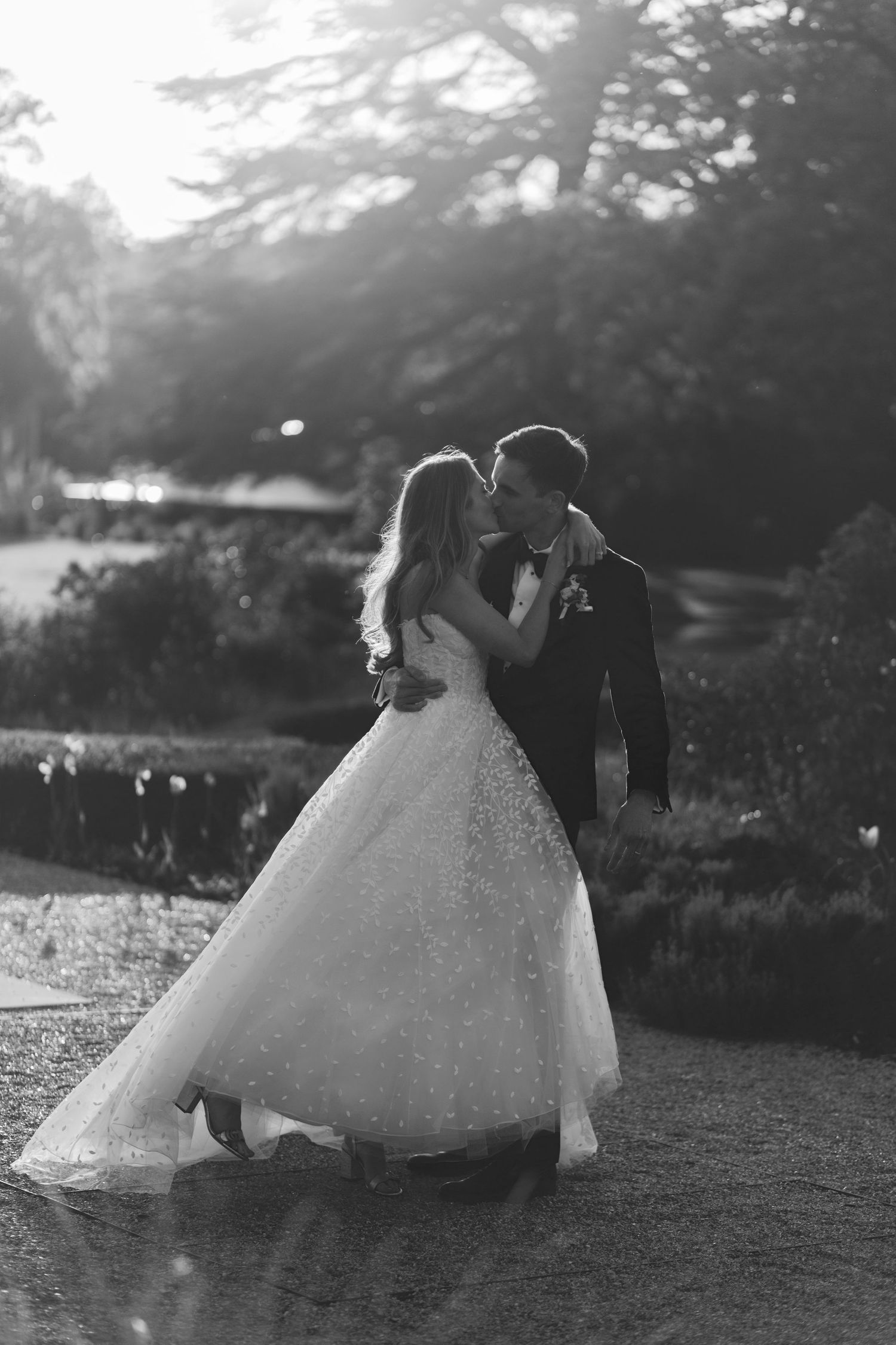Black and white wedding photo of bride and groom sharing a romantic kiss in a lush garden setting.