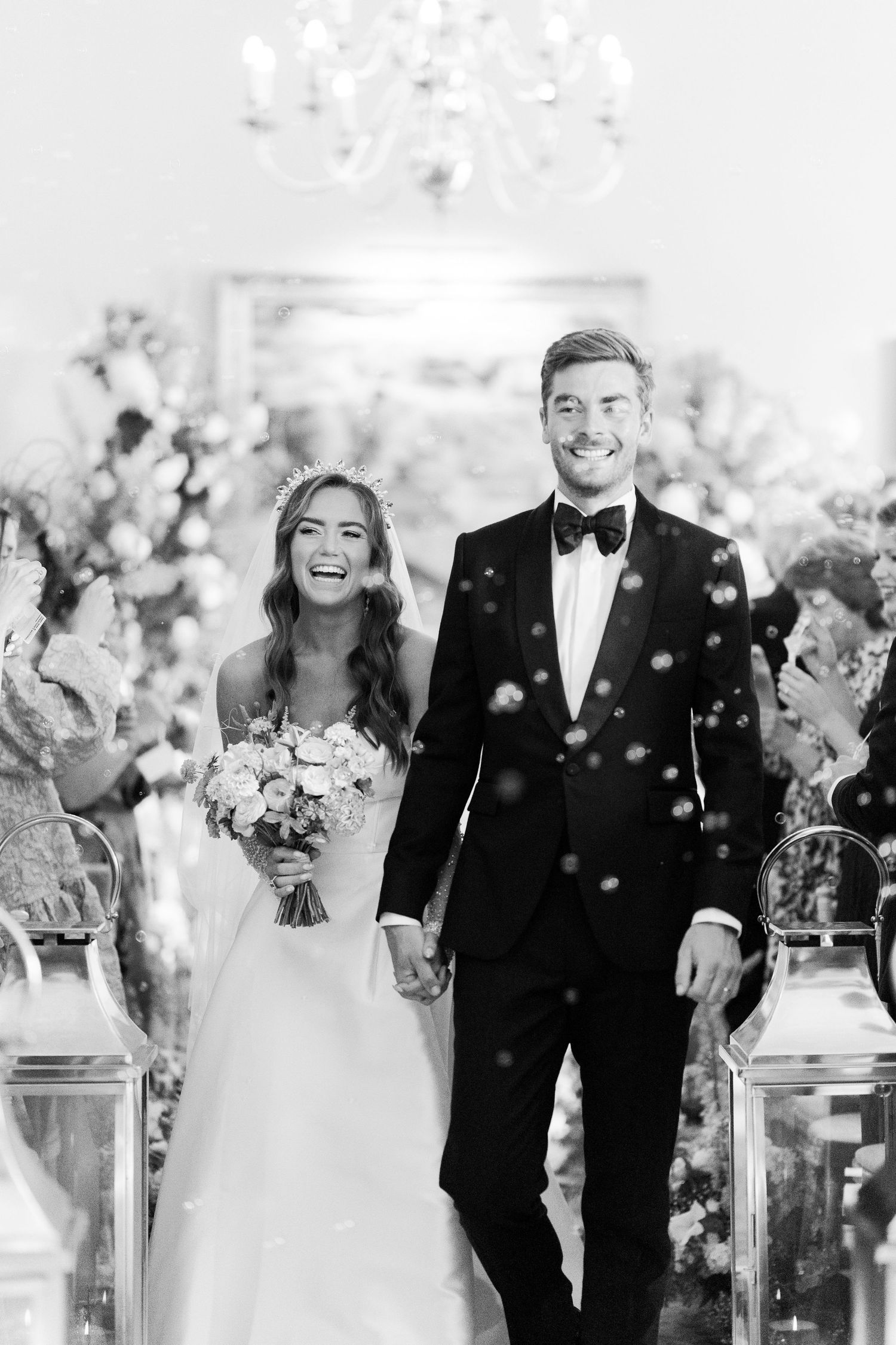 Black and white photo of joyful newlyweds walking down the aisle amid confetti at a chandelier-lit wedding ceremony.