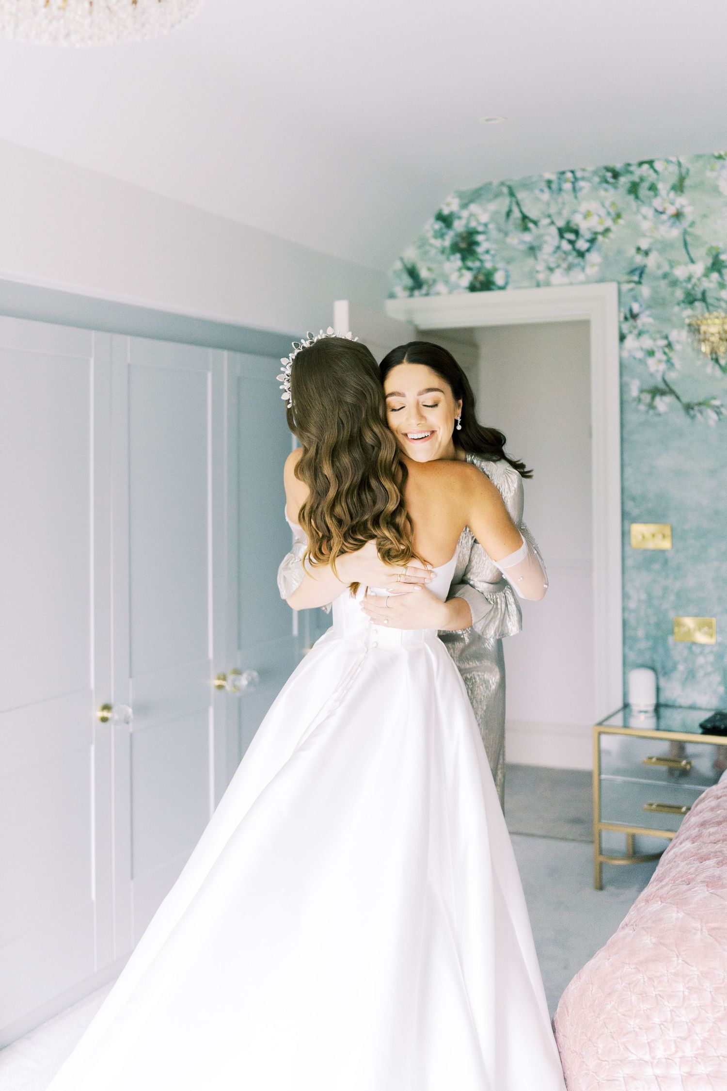 Two women in a bright room, one in a white wedding gown and tiara, sharing a warm embrace while smiling.
