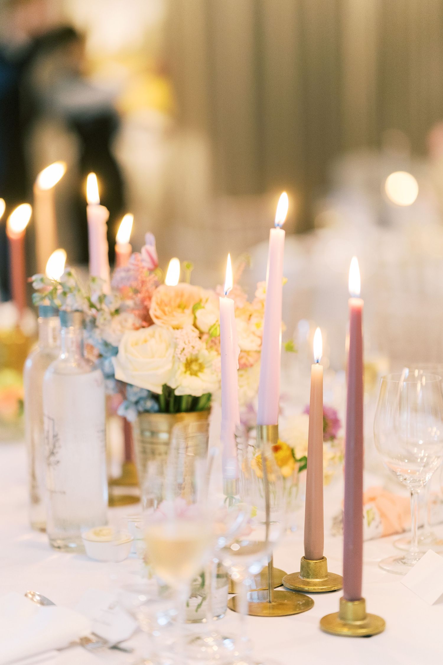 Elegant wedding reception table with lit pink taper candles in gold holders, pastel floral arrangements, and crystal glassware.
