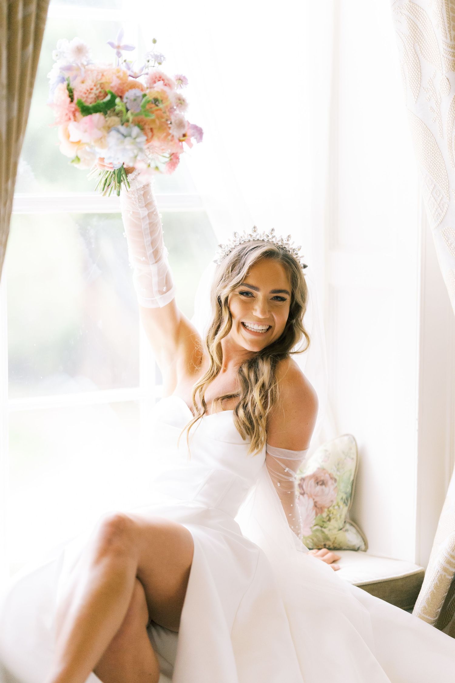 Radiant bride in white strapless gown and tiara raises pastel bouquet while seated by bright window.