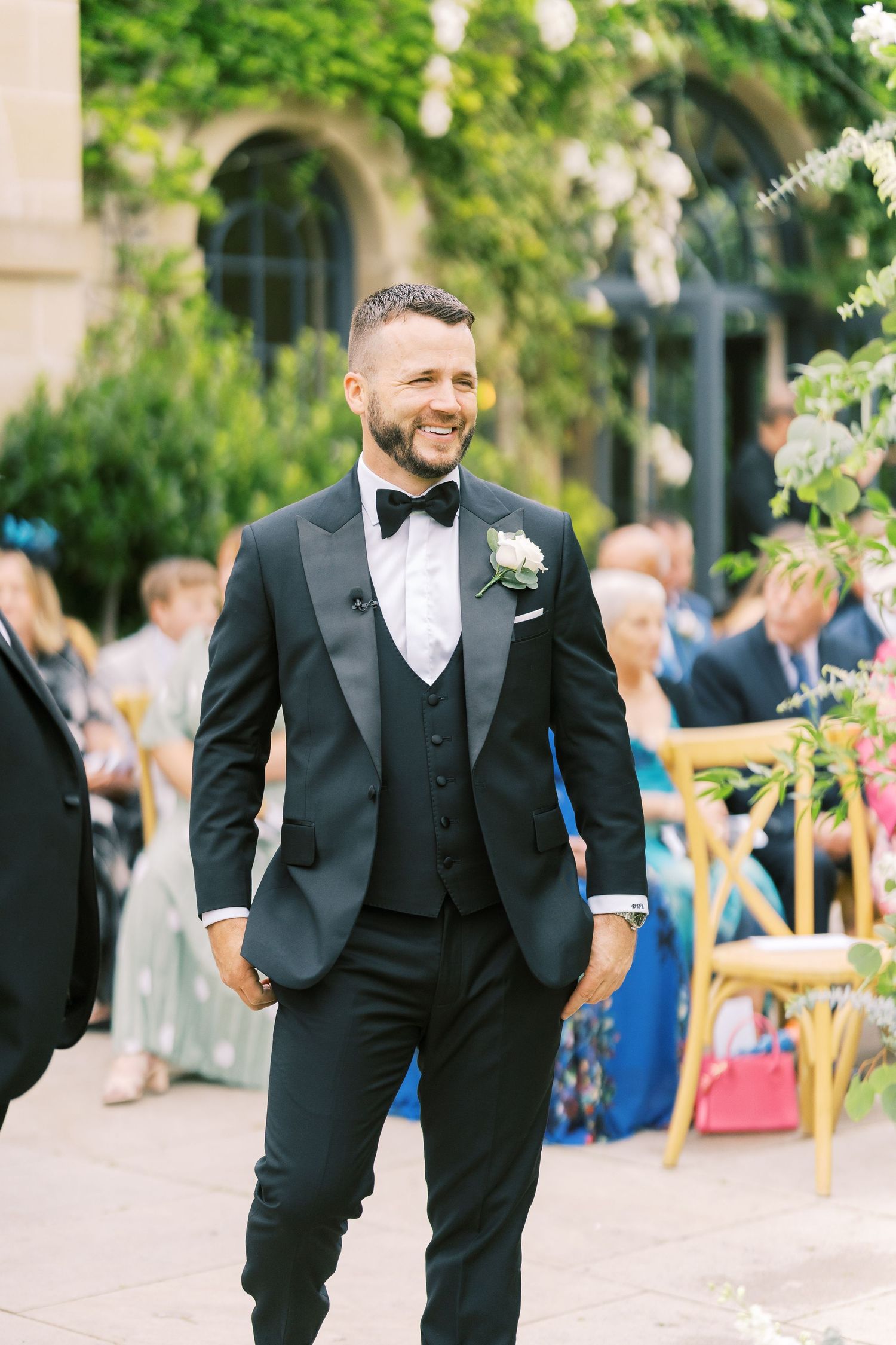Smiling groom in black tuxedo with bow tie and boutonniere walks at outdoor wedding ceremony.