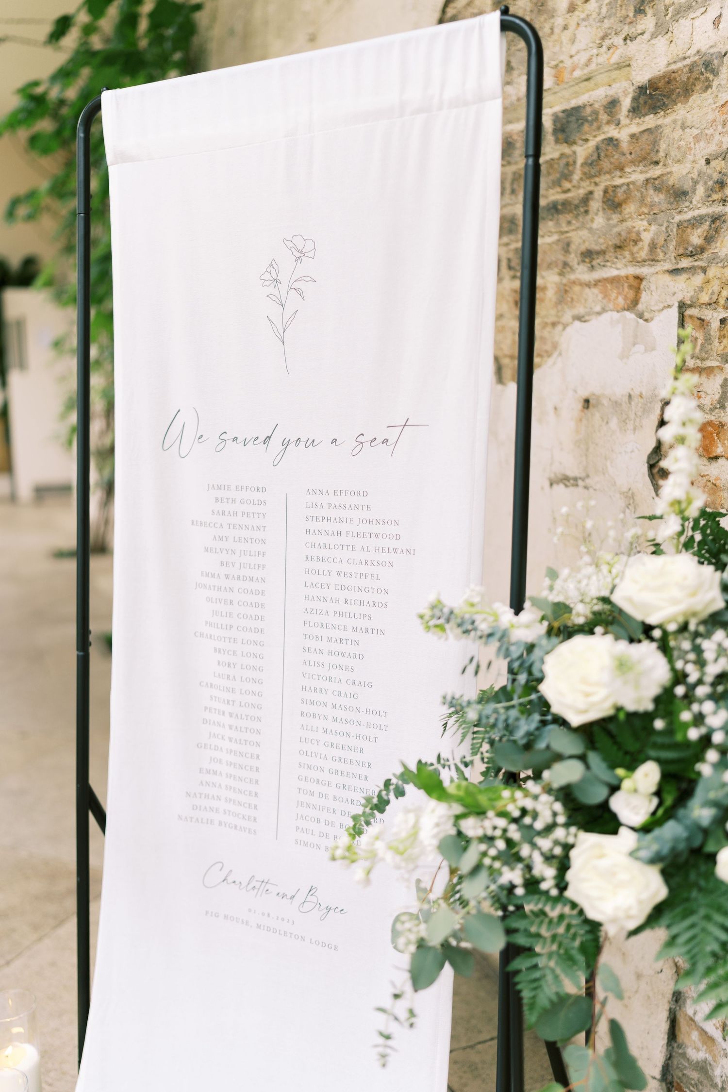 Elegant wedding seating chart on white fabric with floral illustration, displayed on black stand with white rose arrangements.