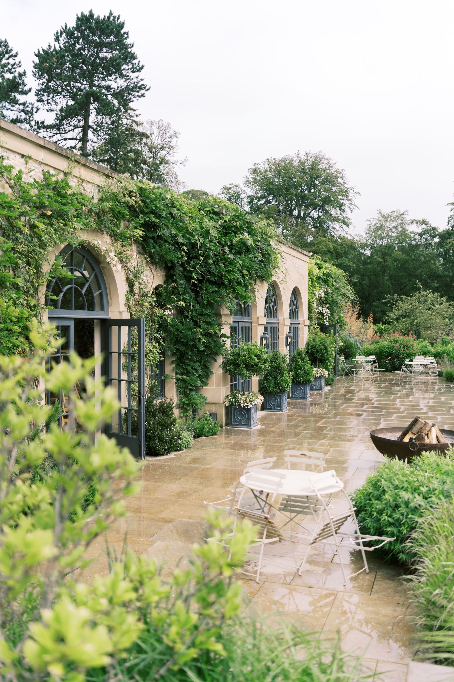 Ivy-covered stone orangery with arched windows, wet patio, white bistro furniture, and lush garden surroundings.