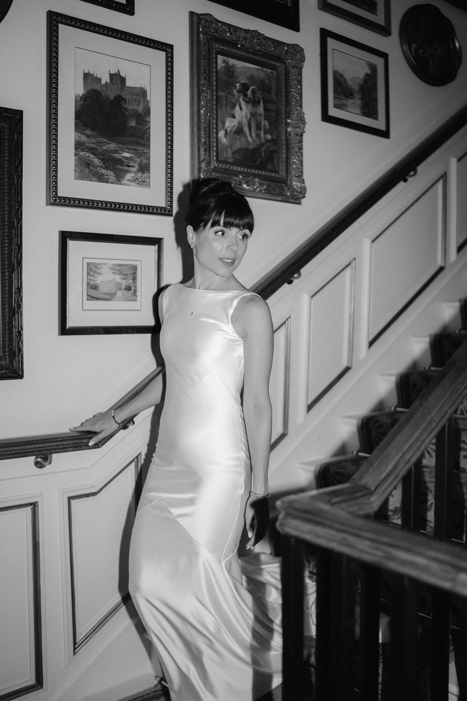 Bride in sleek white satin gown posing on staircase surrounded by framed artwork in black and white.