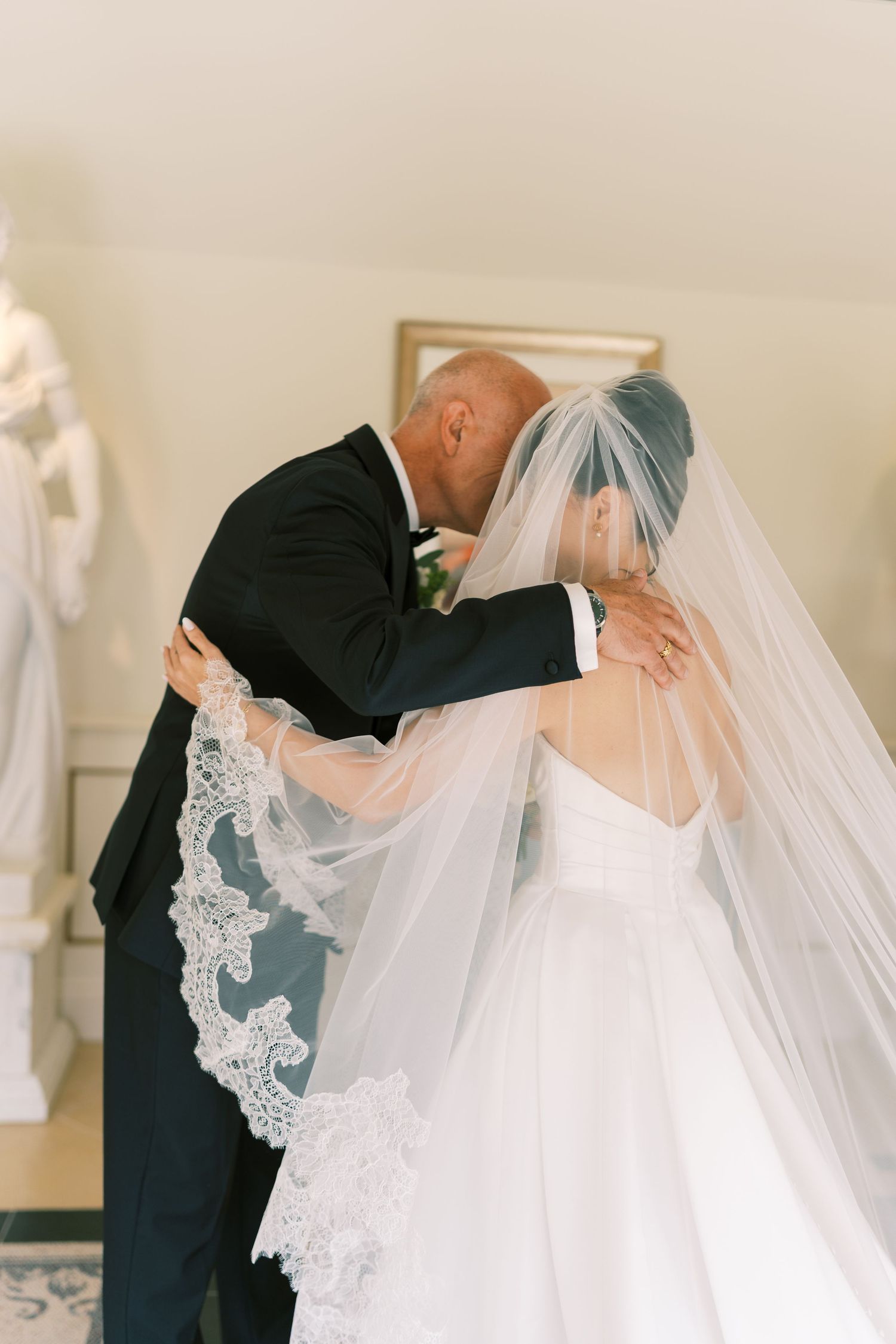 Bride and groom share an intimate embrace, her lace-trimmed veil cascading down her back in elegant wedding attire.