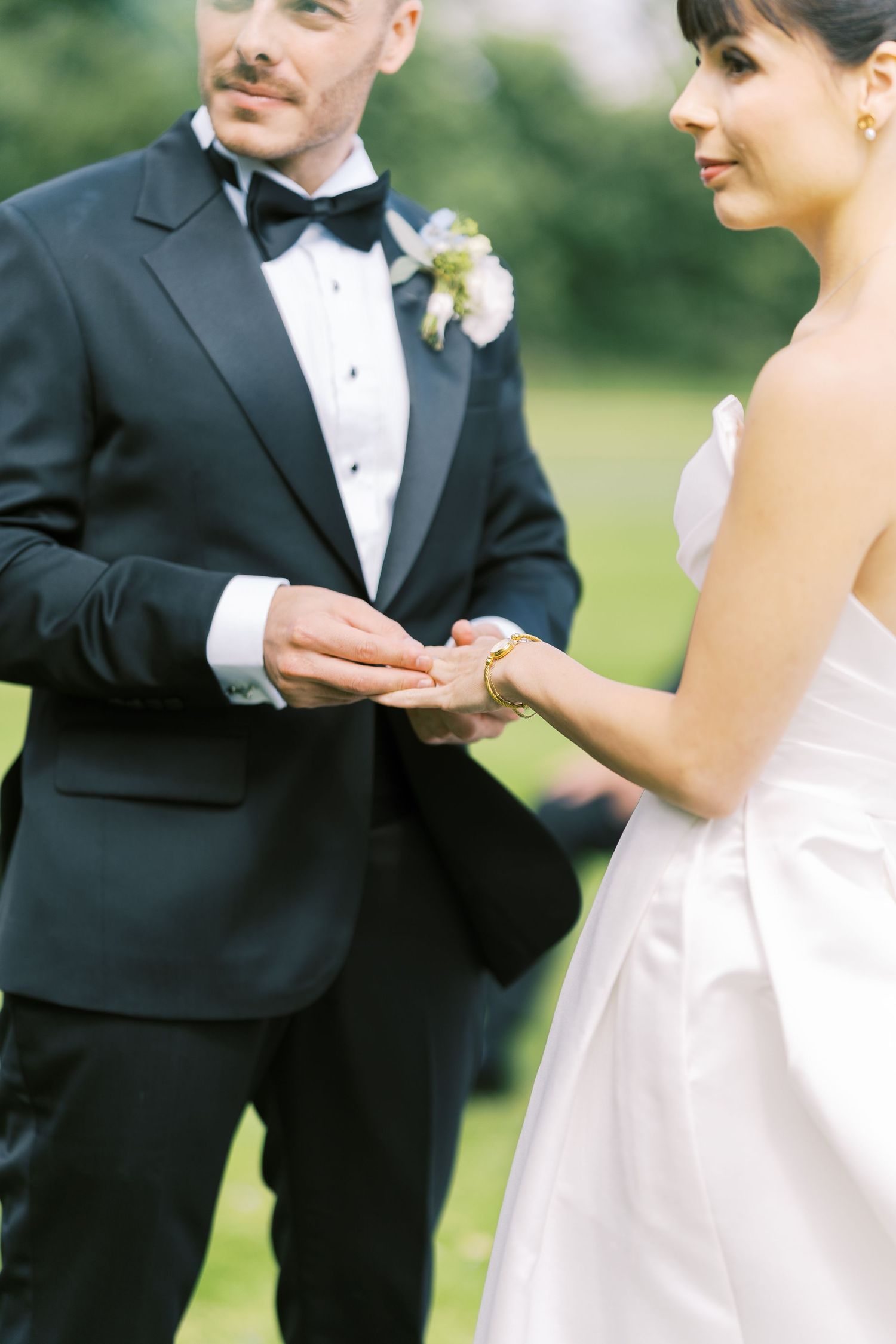Groom in black tuxedo places wedding ring on brides finger during outdoor ceremony.