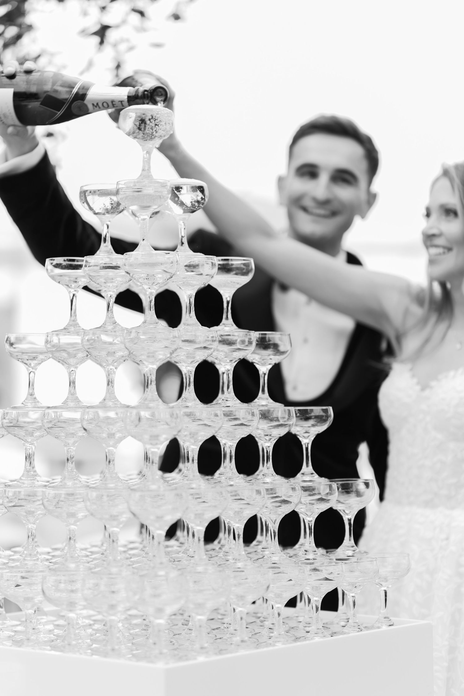 Black and white photo of newlyweds pouring champagne into a pyramid of glasses at their wedding reception.