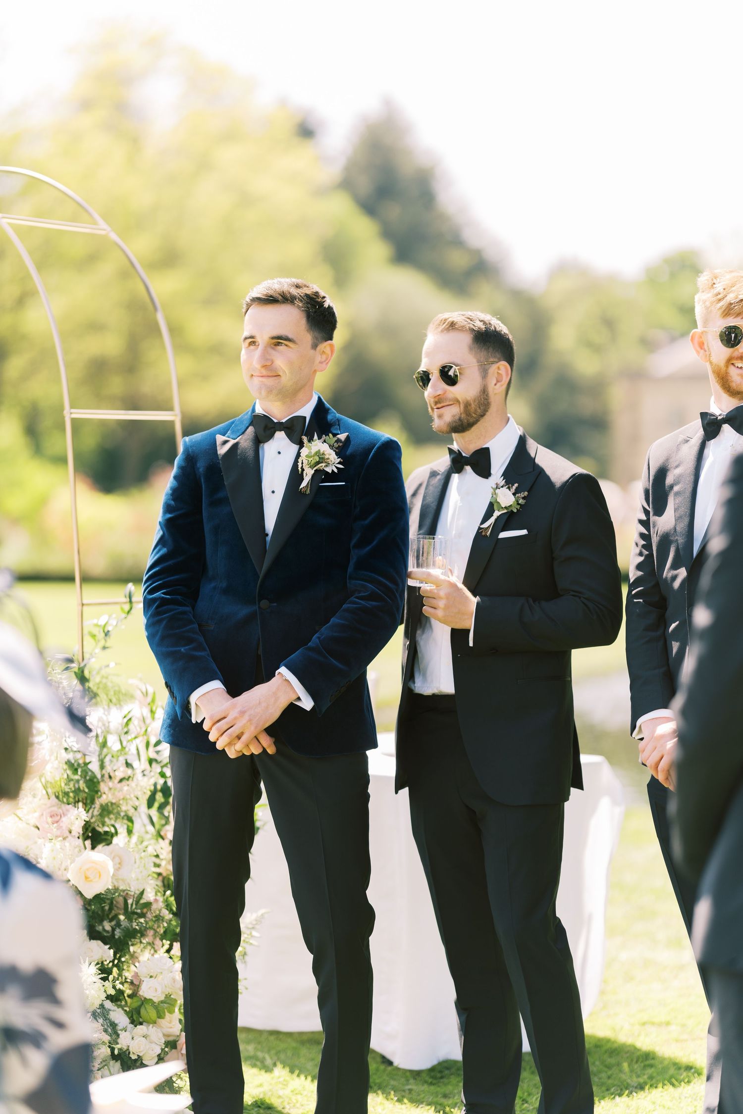 Groom in navy velvet tuxedo stands at outdoor wedding ceremony altar with groomsmen.