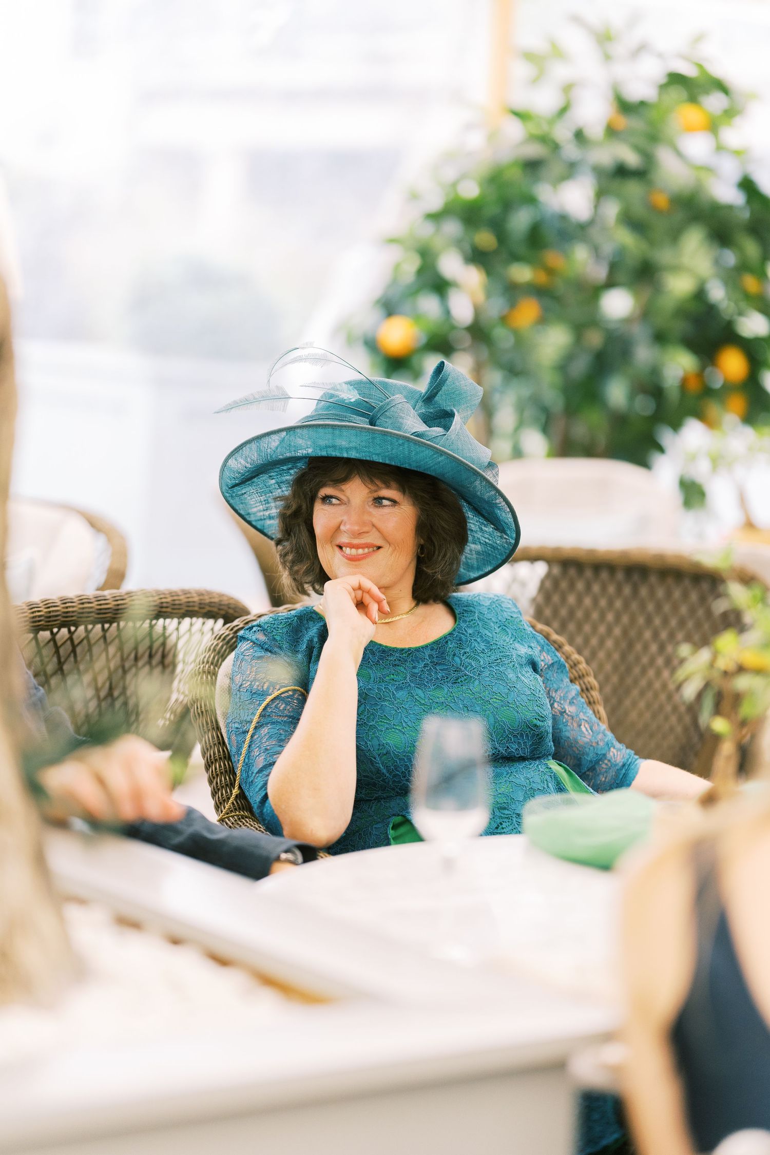 Smiling woman in teal lace dress and matching wide-brim hat seated at elegant wedding reception table.