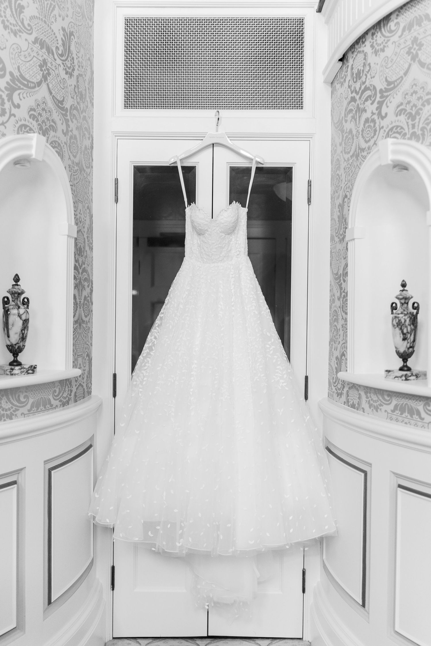 Black and white photo of an elegant strapless ball gown wedding dress hanging on a door in a ornate room.