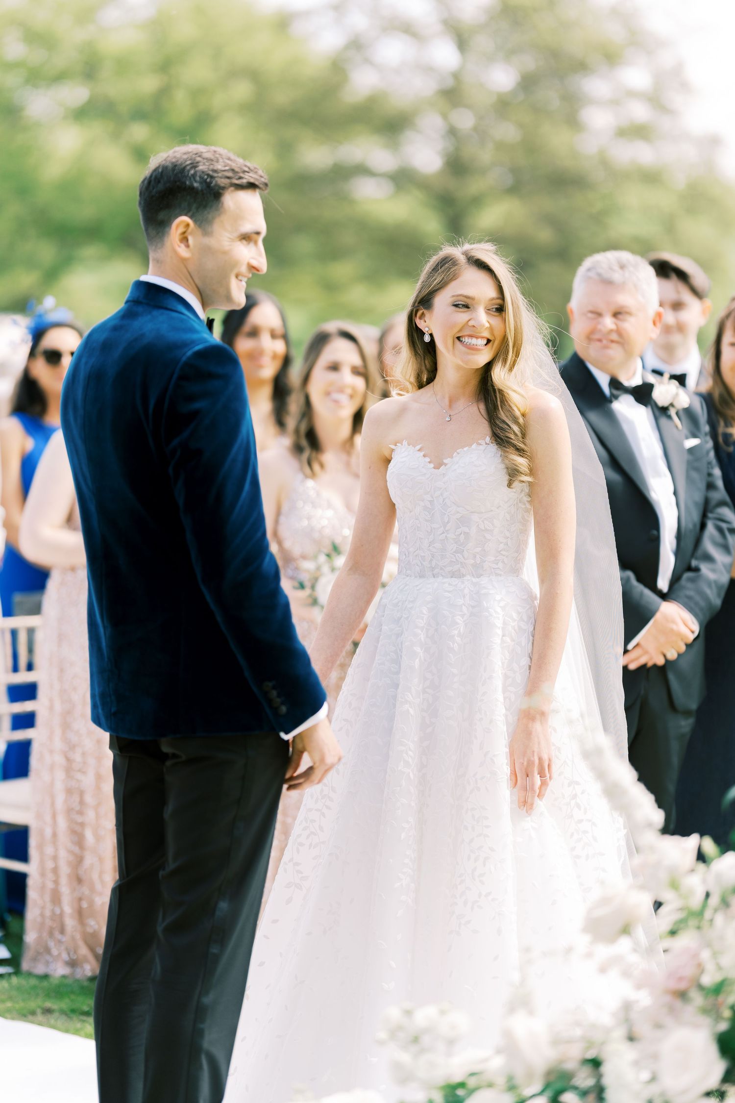 Bride in white lace gown smiles at groom during outdoor wedding ceremony.