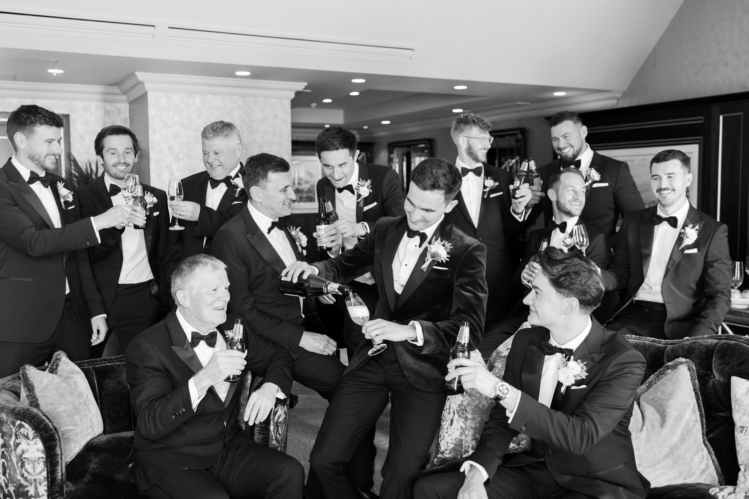 Groomsmen in tuxedos celebrate with champagne in a black and white wedding photo.
