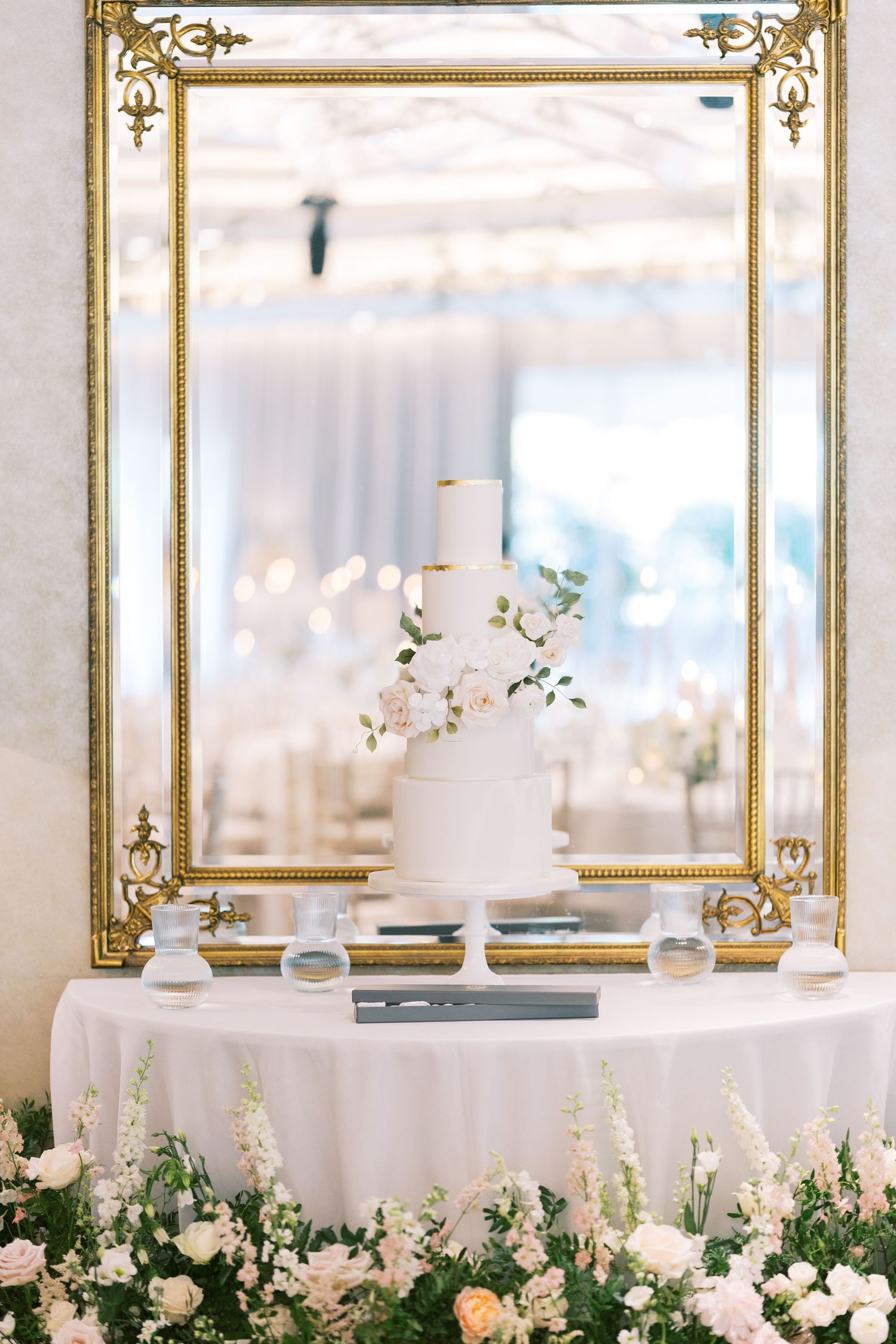Elegant three-tier white wedding cake with floral decor on a draped table, framed by a gold mirror and white roses.
