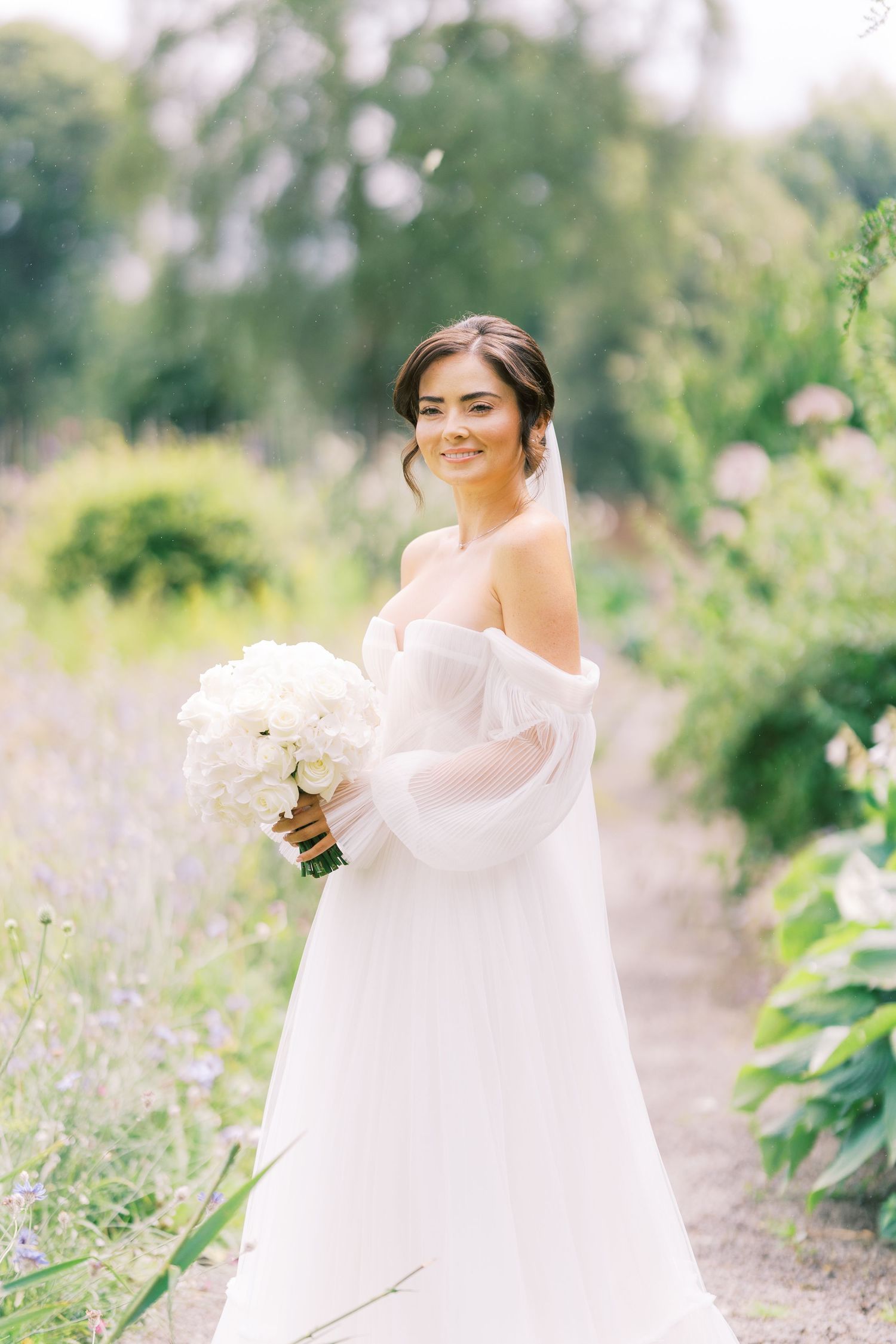Smiling bride in off-shoulder white gown holds hydrangea bouquet in lush garden setting.
