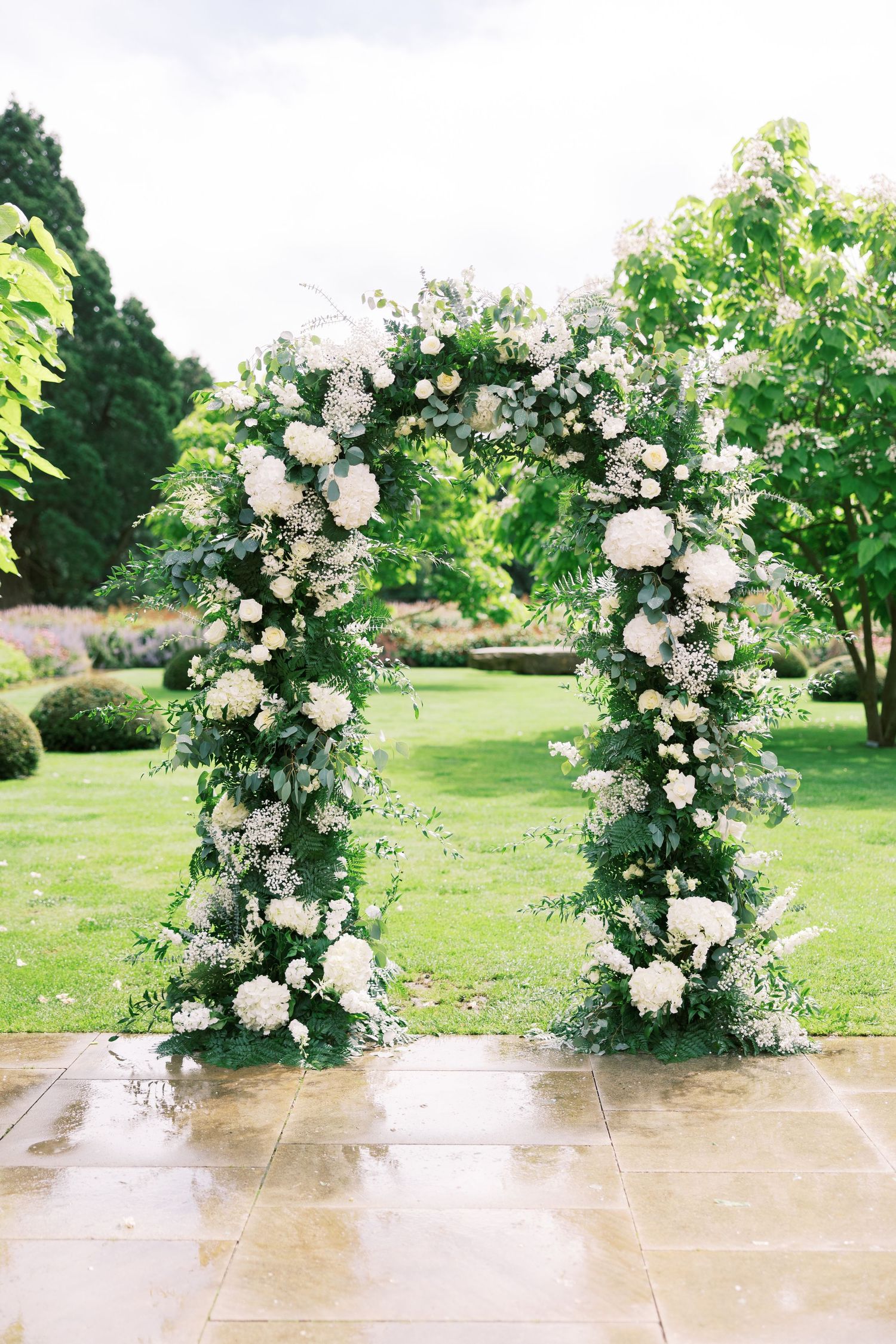 Elegant outdoor wedding arch adorned with lush white hydrangeas and greenery, set against a manicured garden backdrop.