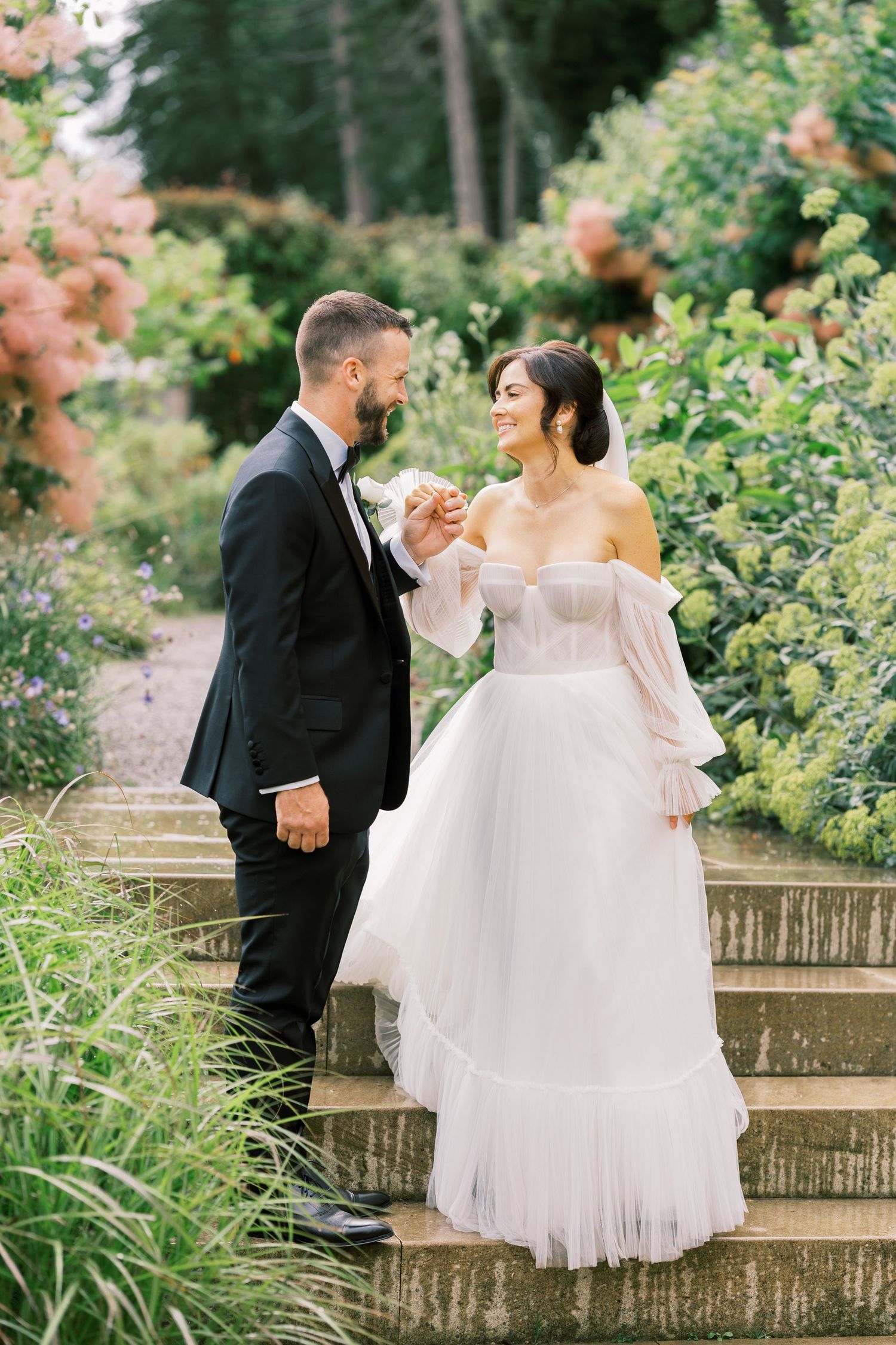 Bride and groom hold hands on garden steps, she wears off-shoulder tulle gown, he wears black tuxedo.