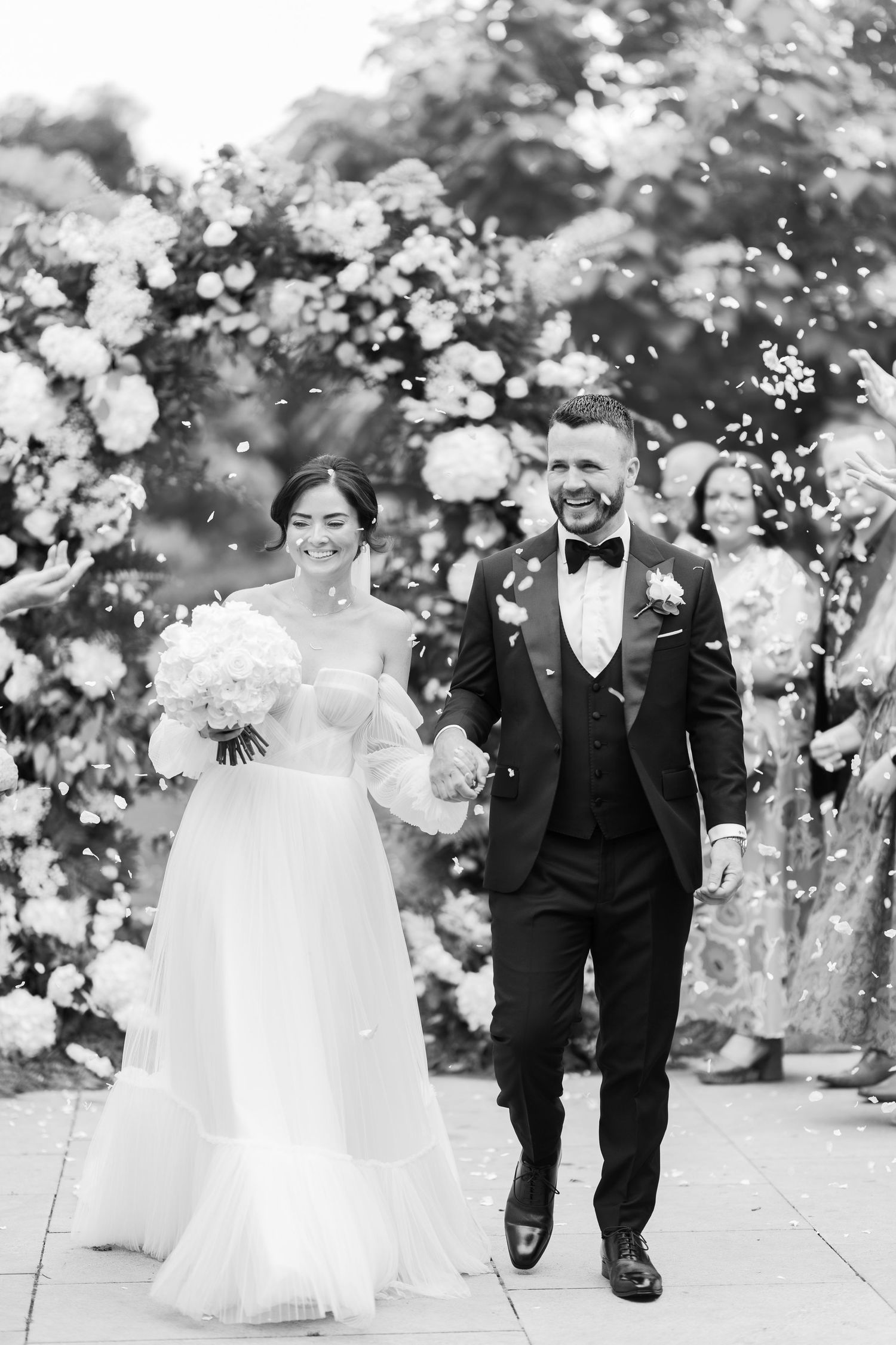 Black and white photo of a smiling bride and groom walking hand in hand through confetti after their wedding ceremony.