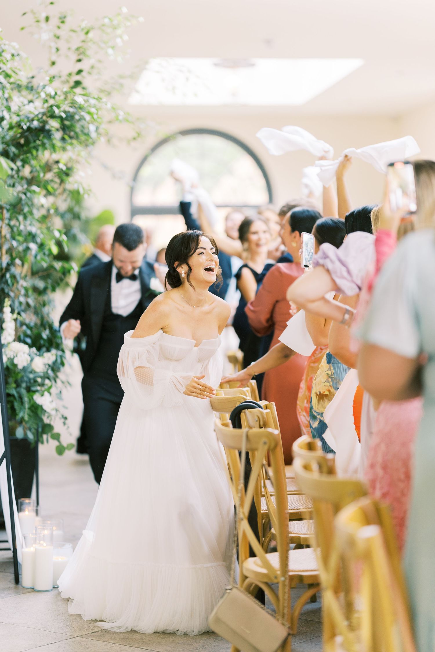 Joyful bride in off-shoulder white gown walks down aisle as cheering wedding guests wave napkins in celebration.