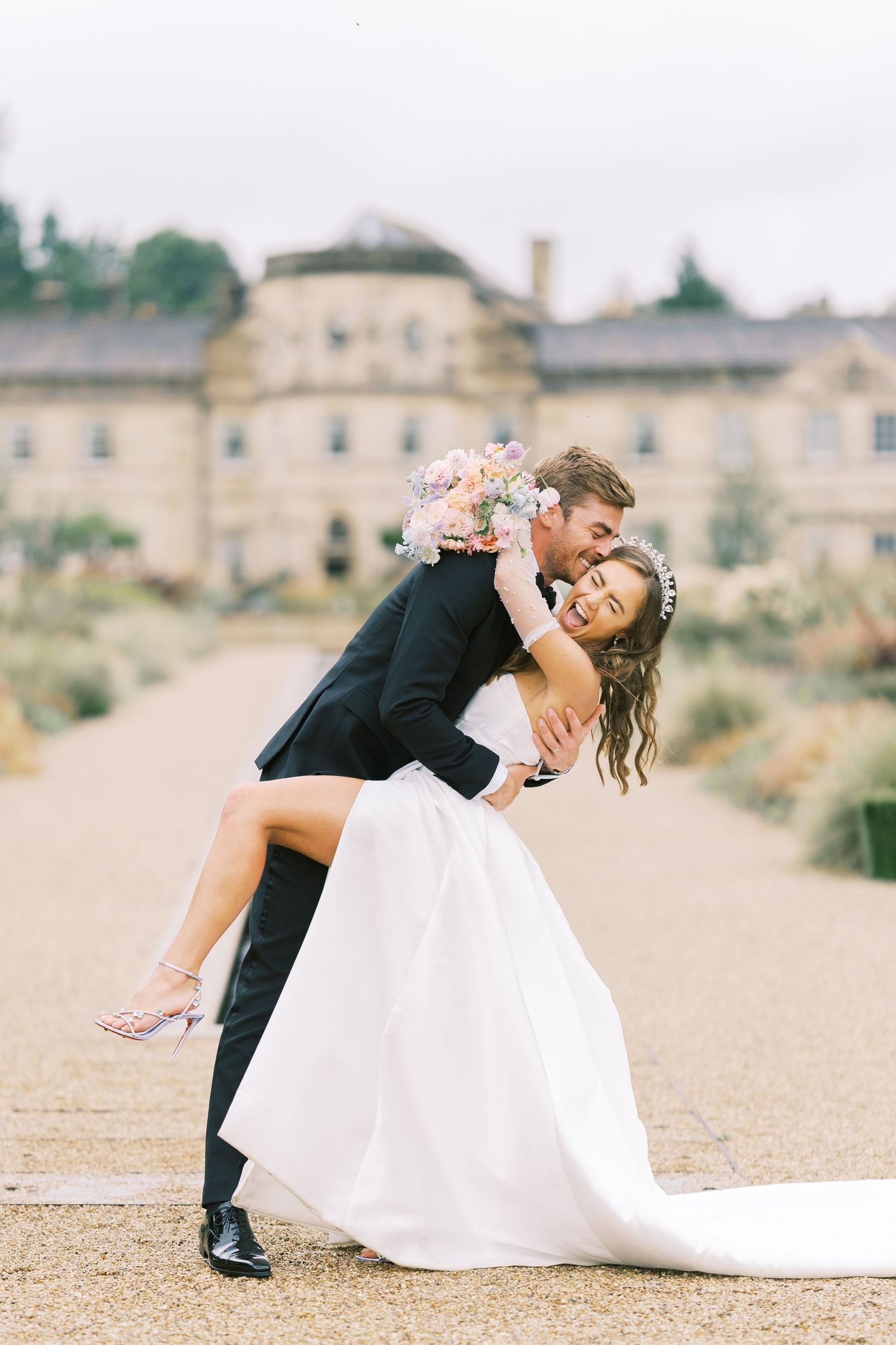 Groom dipping bride in white gown and floral bouquet outside elegant stone manor on their wedding day.