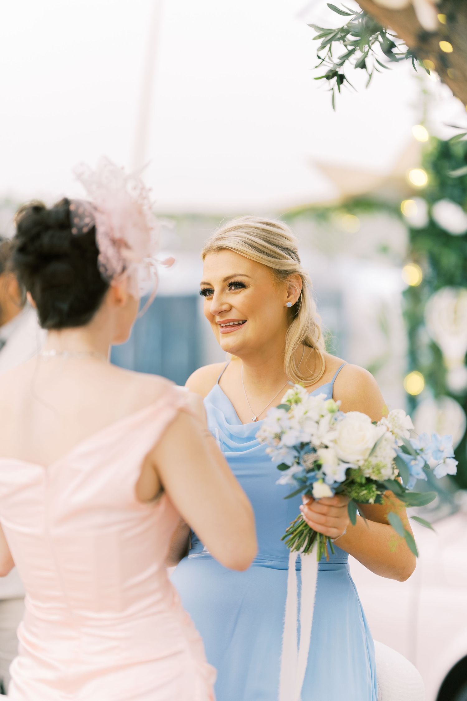 Smiling blonde bridesmaid in blue dress holds white bouquet while conversing at elegant wedding reception.