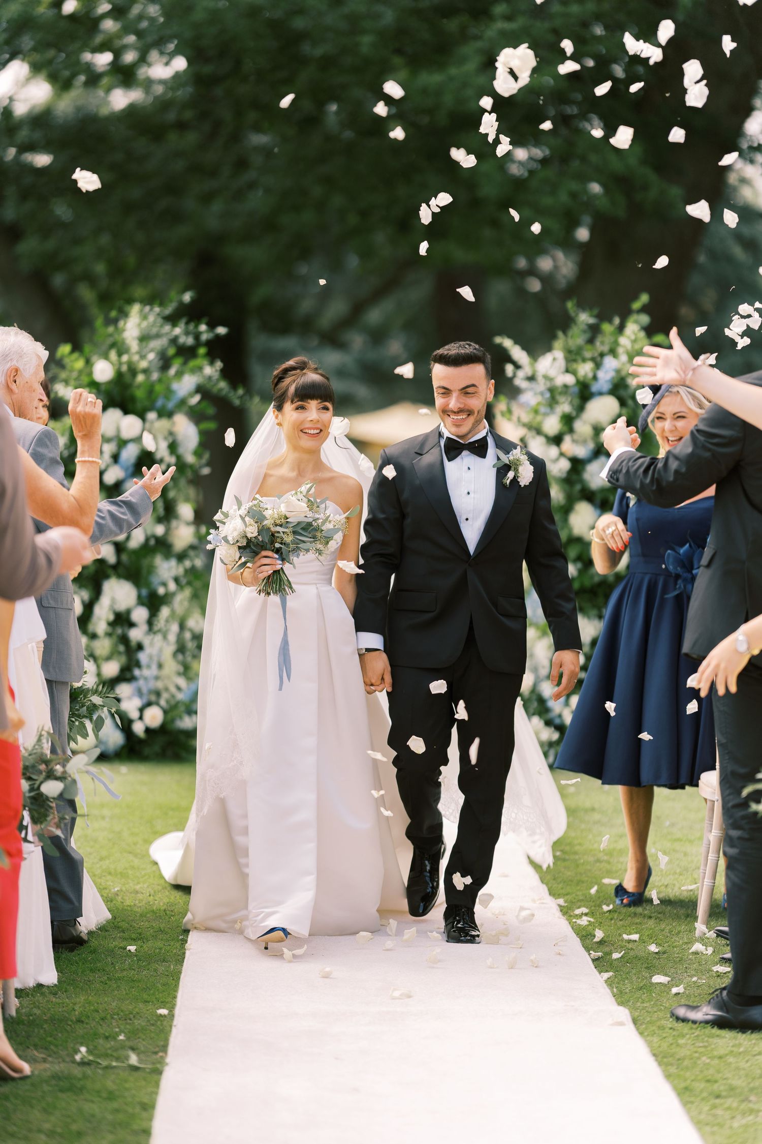 Joyful bride and groom walk down aisle as guests toss white flower petals at outdoor garden wedding.