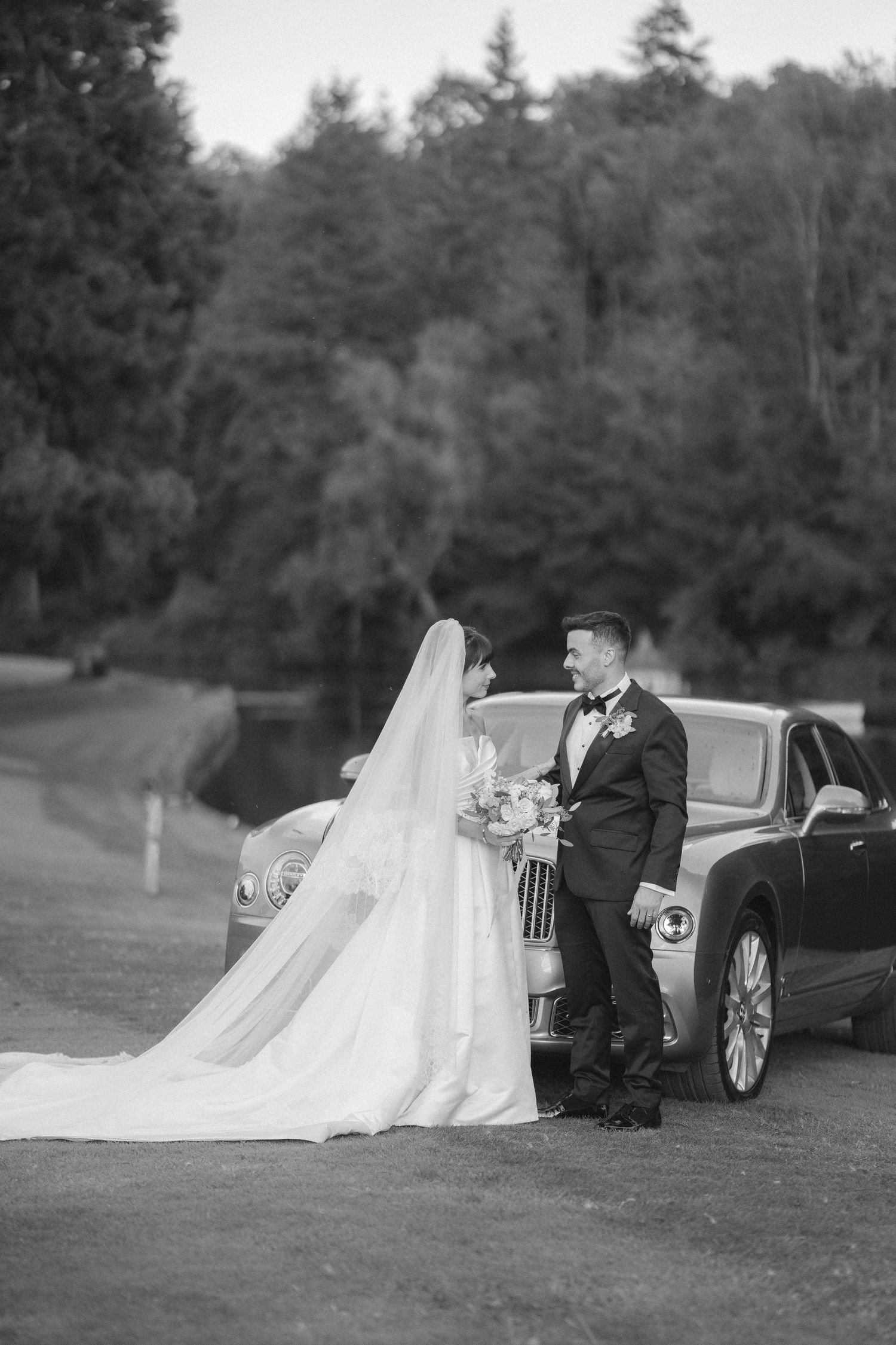 Black and white photo of bride and groom posing by a luxury car with lush trees in the background.