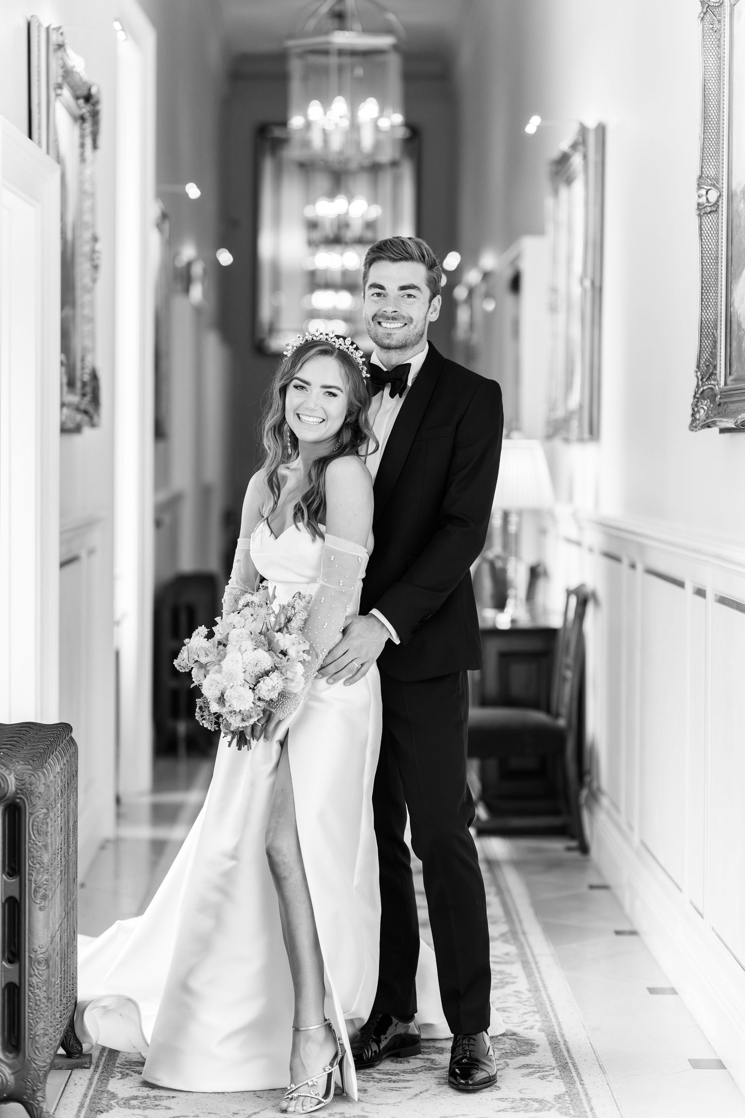 Black and white photo of a smiling bride and groom posing in an elegant hallway with chandelier.