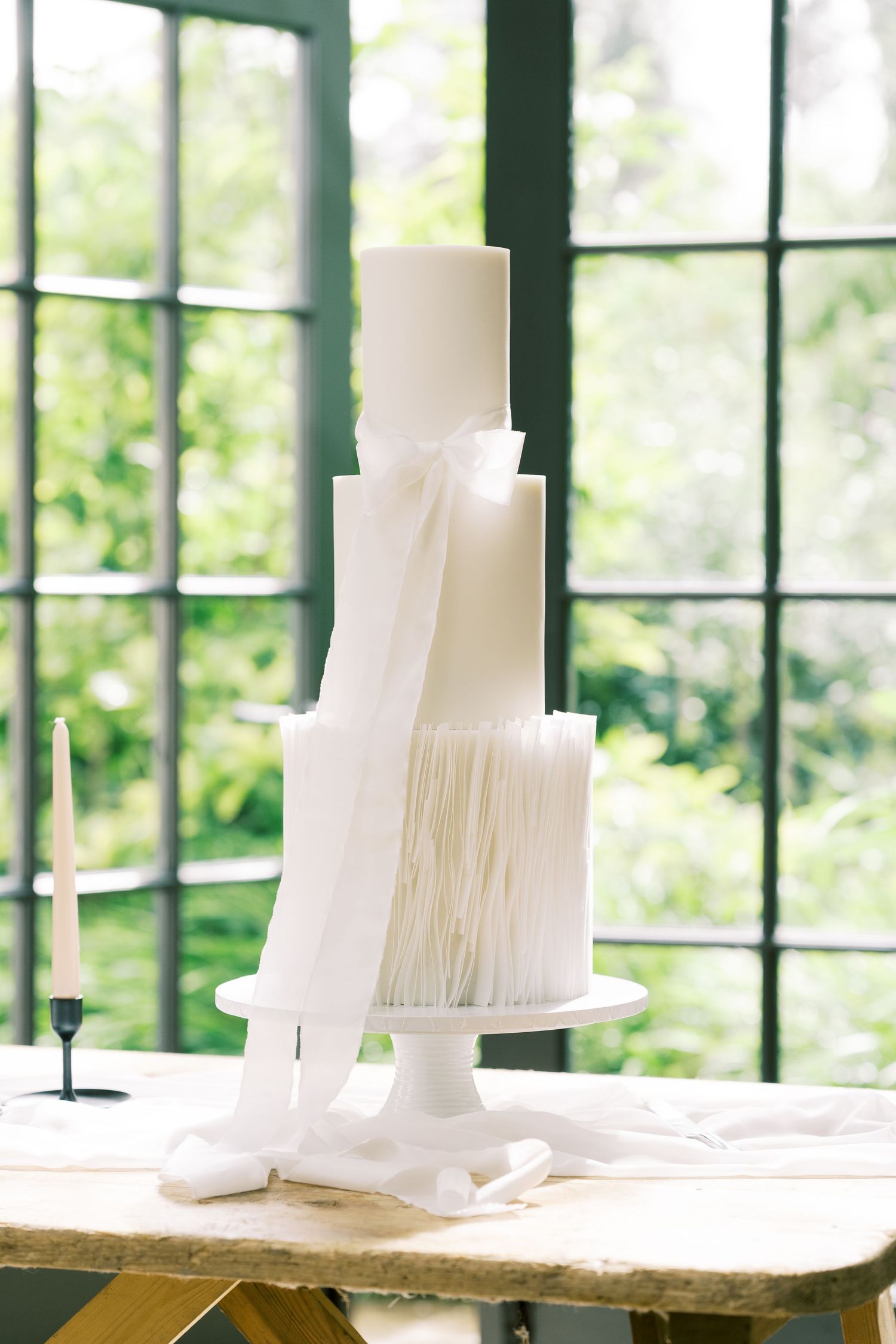 Elegant three-tier white wedding cake with ribbon and fringe details on a cake stand by a window.