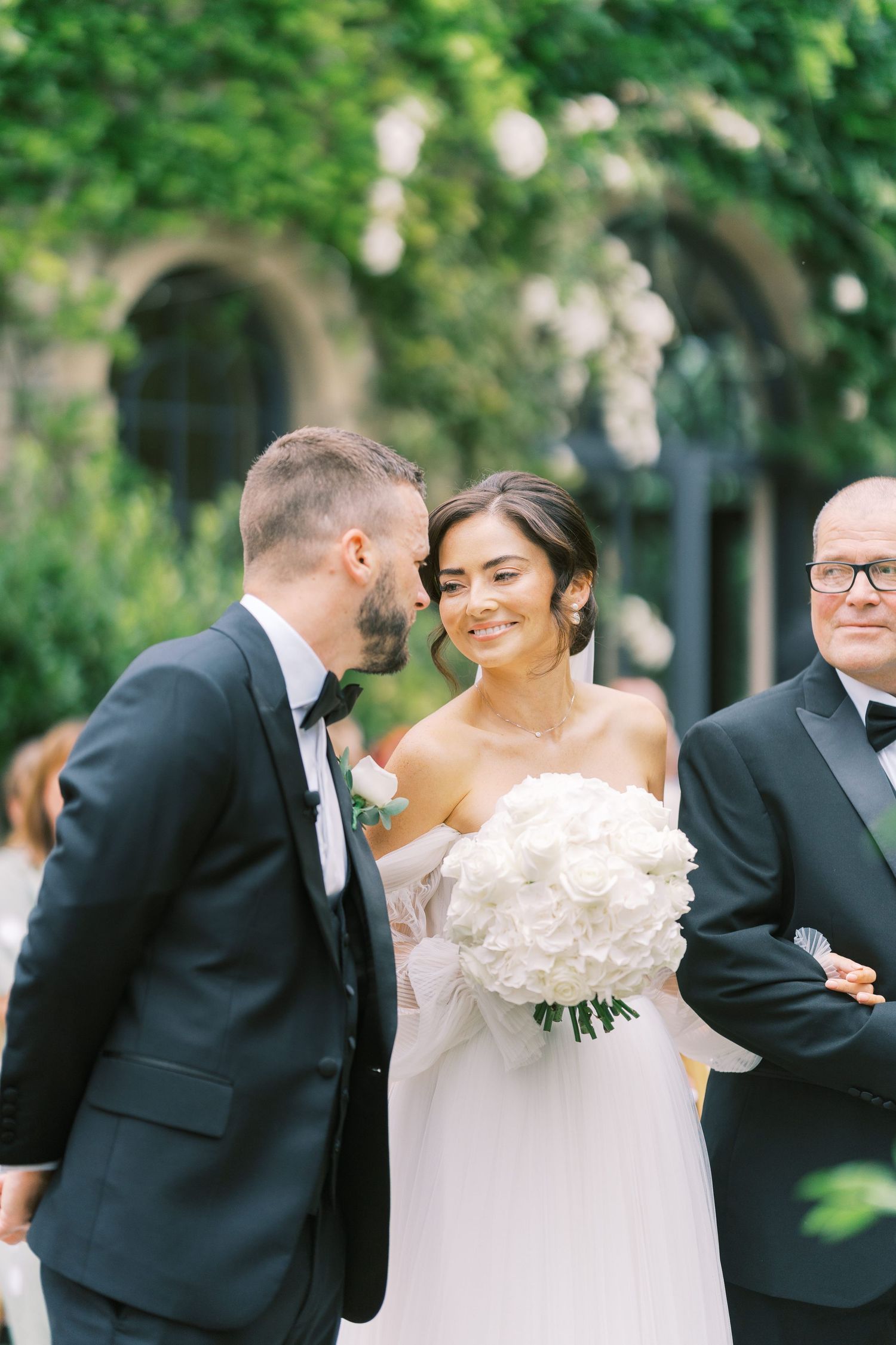 Bride holding white rose bouquet smiles at groom in black tuxedo during outdoor wedding ceremony.