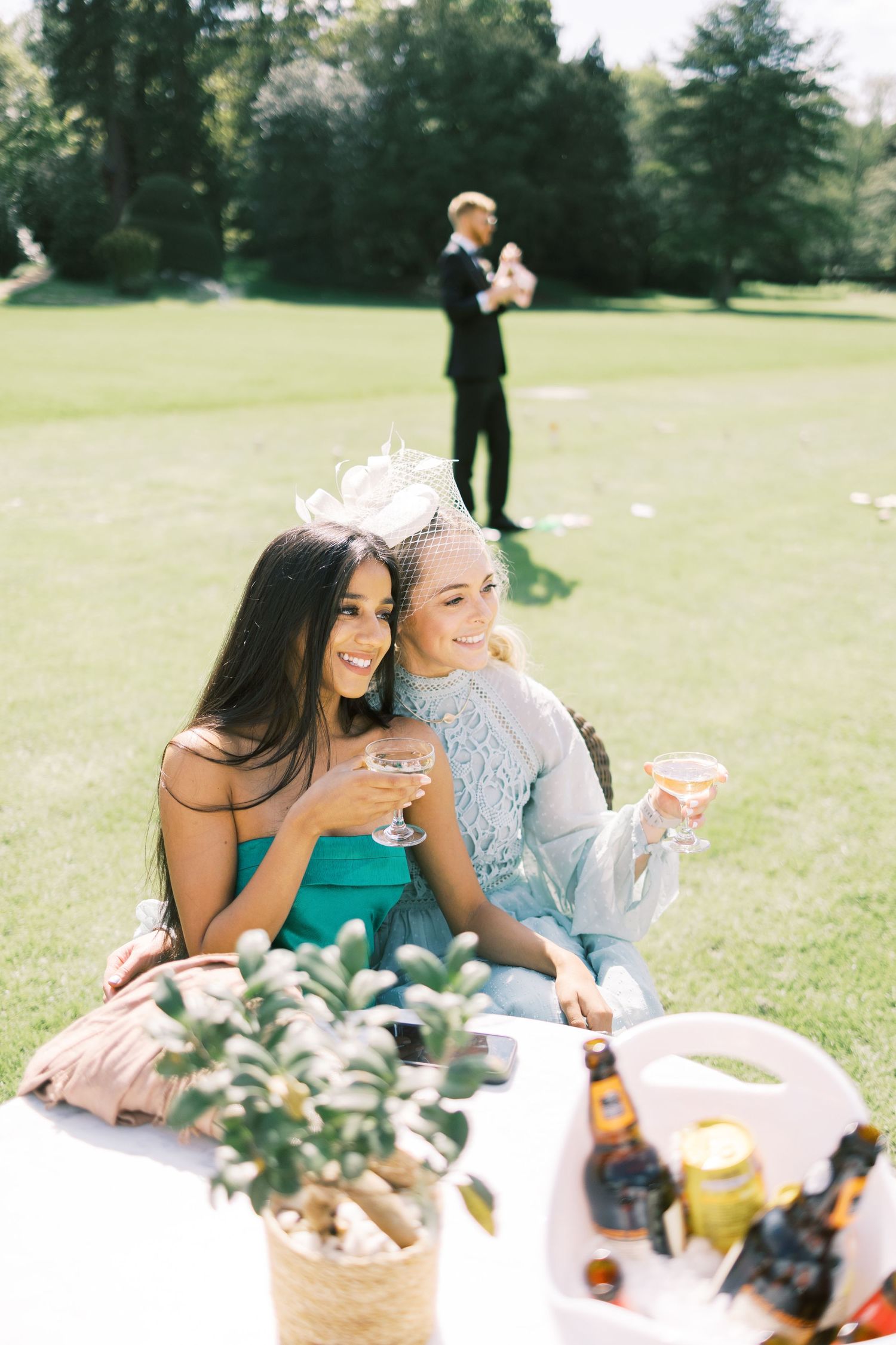 Two elegantly dressed women wearing fascinators enjoy cocktails outdoors at a sunny garden party event.