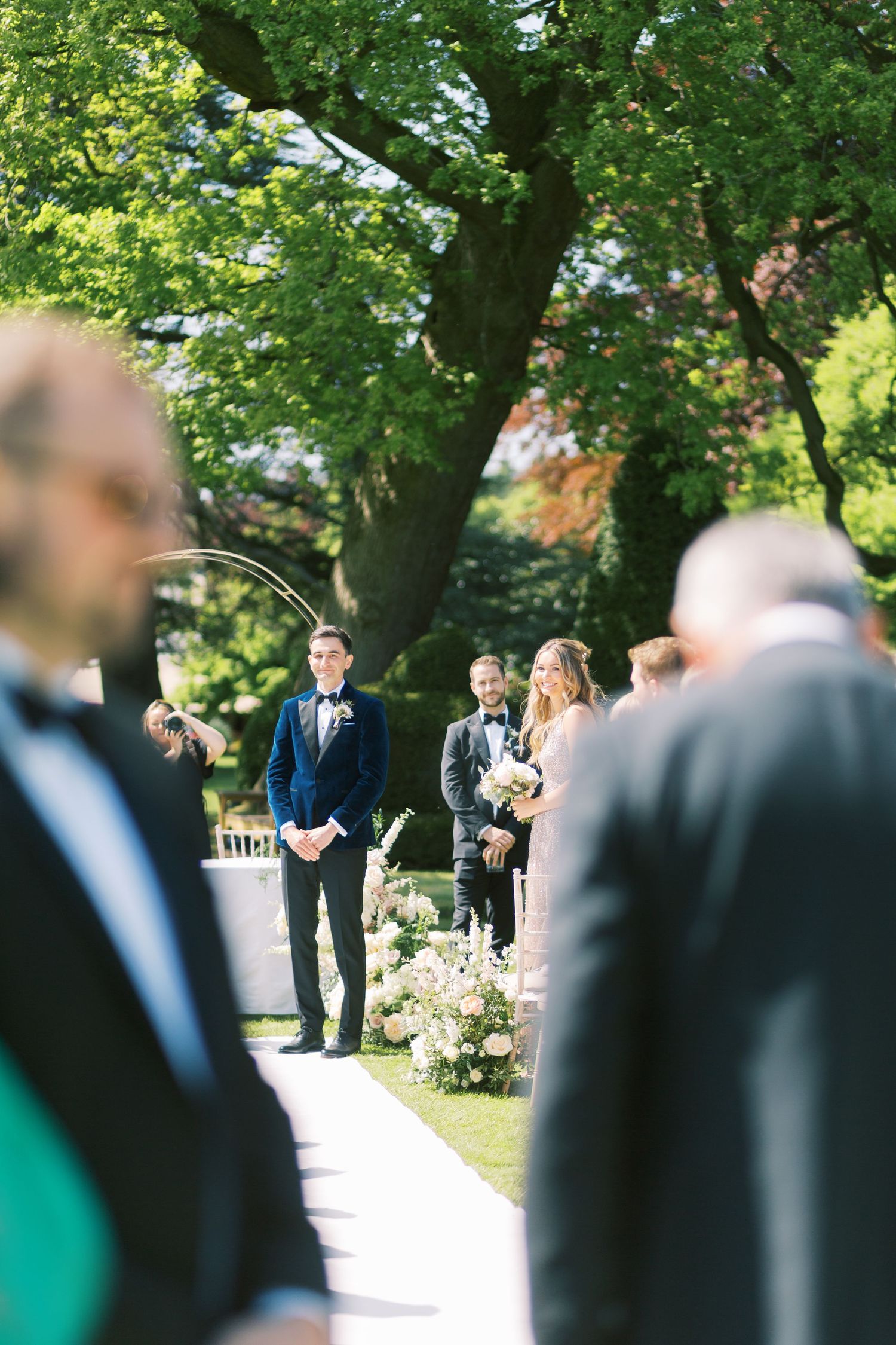 Groom in navy velvet tuxedo watches bride walk down outdoor aisle lined with white floral arrangements.