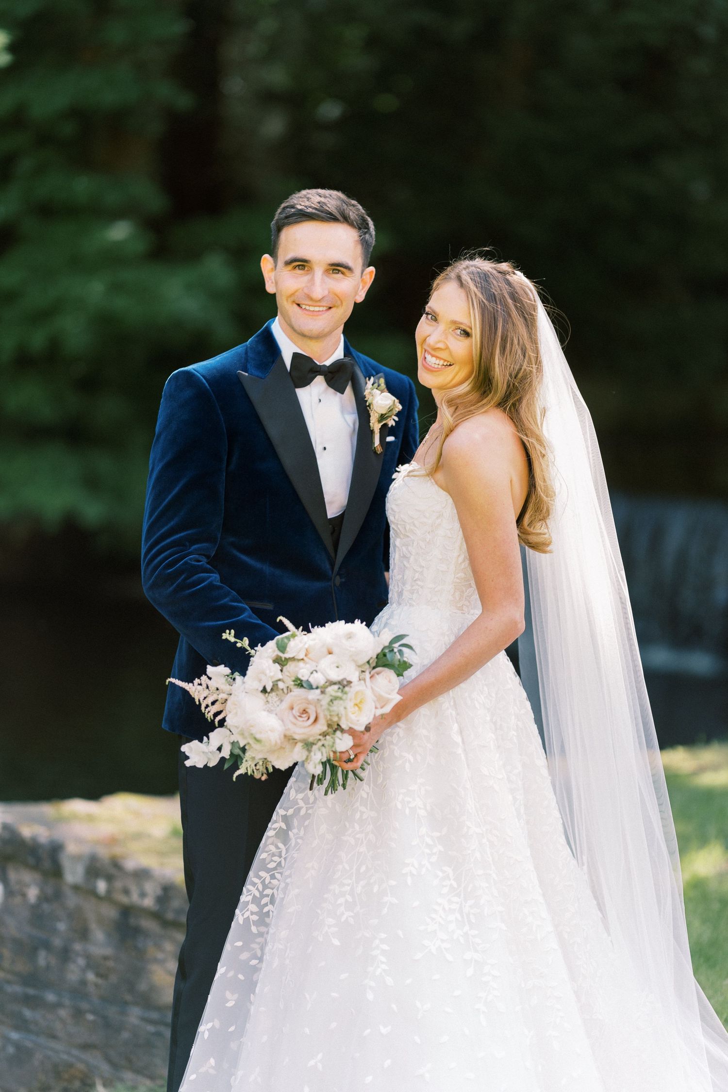 A smiling bride in a lace gown holding a white bouquet stands with a groom in a navy velvet tuxedo outdoors.