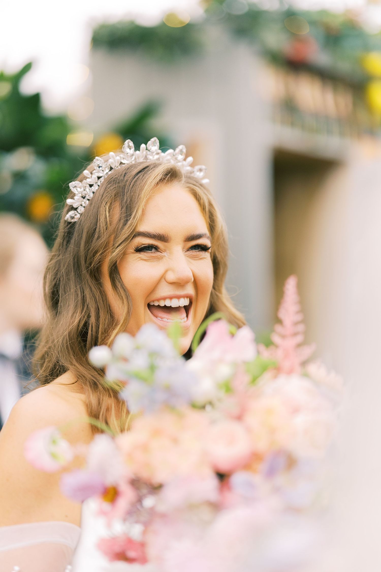 Laughing bride wearing a crystal tiara holds a pastel bouquet of pink and white flowers on her wedding day.