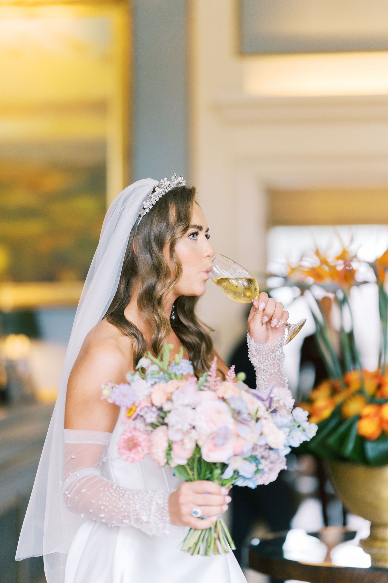 Bride in white gown and tiara sips champagne while holding a pastel floral bouquet indoors.