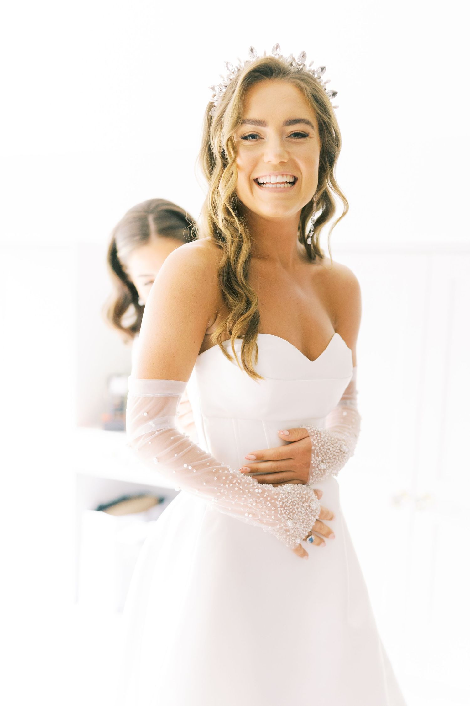 Smiling bride in a strapless white gown with pearl gloves and a sparkling tiara poses in a sunlit room.