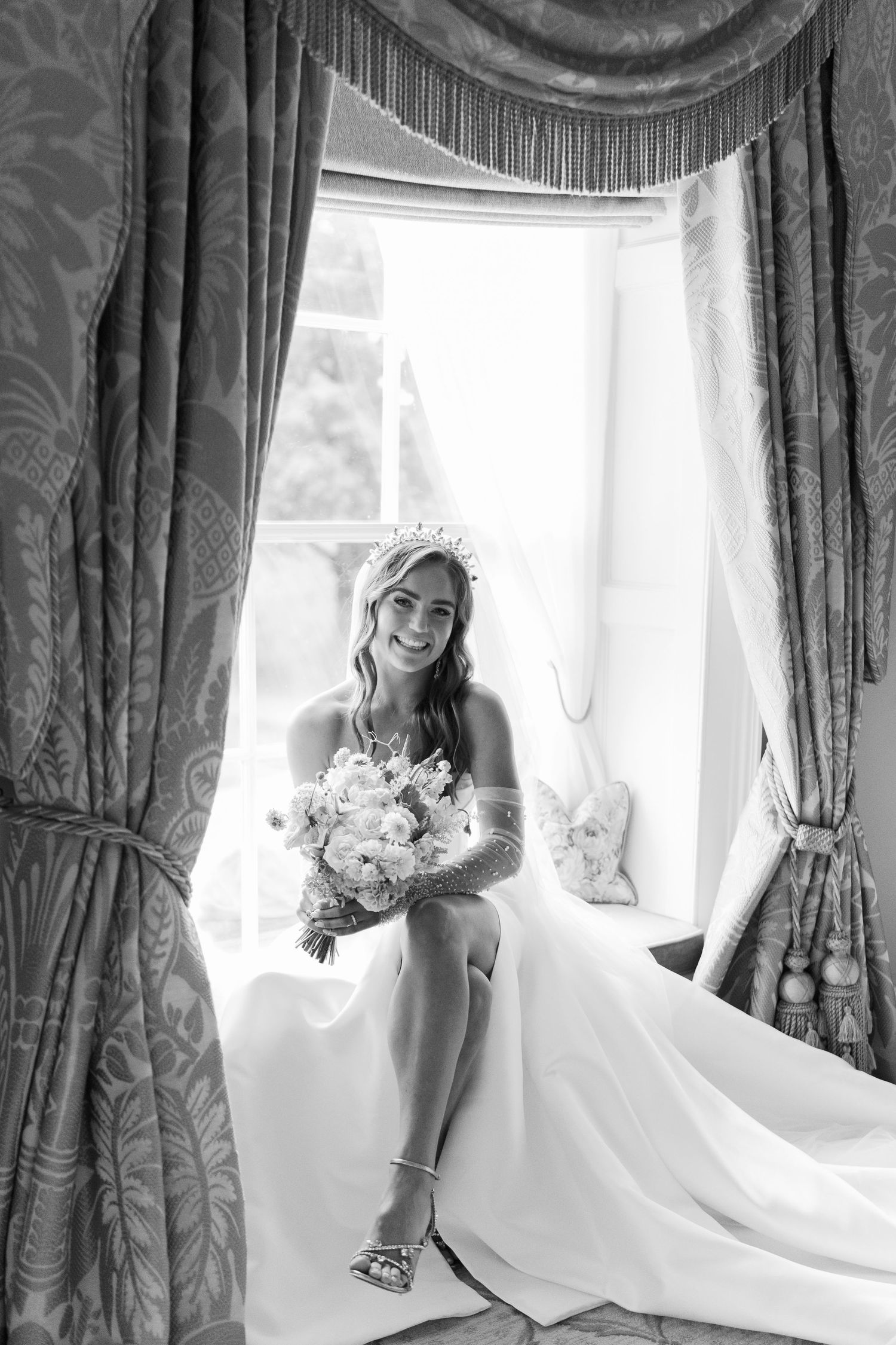 Black and white photo of a smiling bride in a white gown and tiara sitting by a window holding a bouquet.
