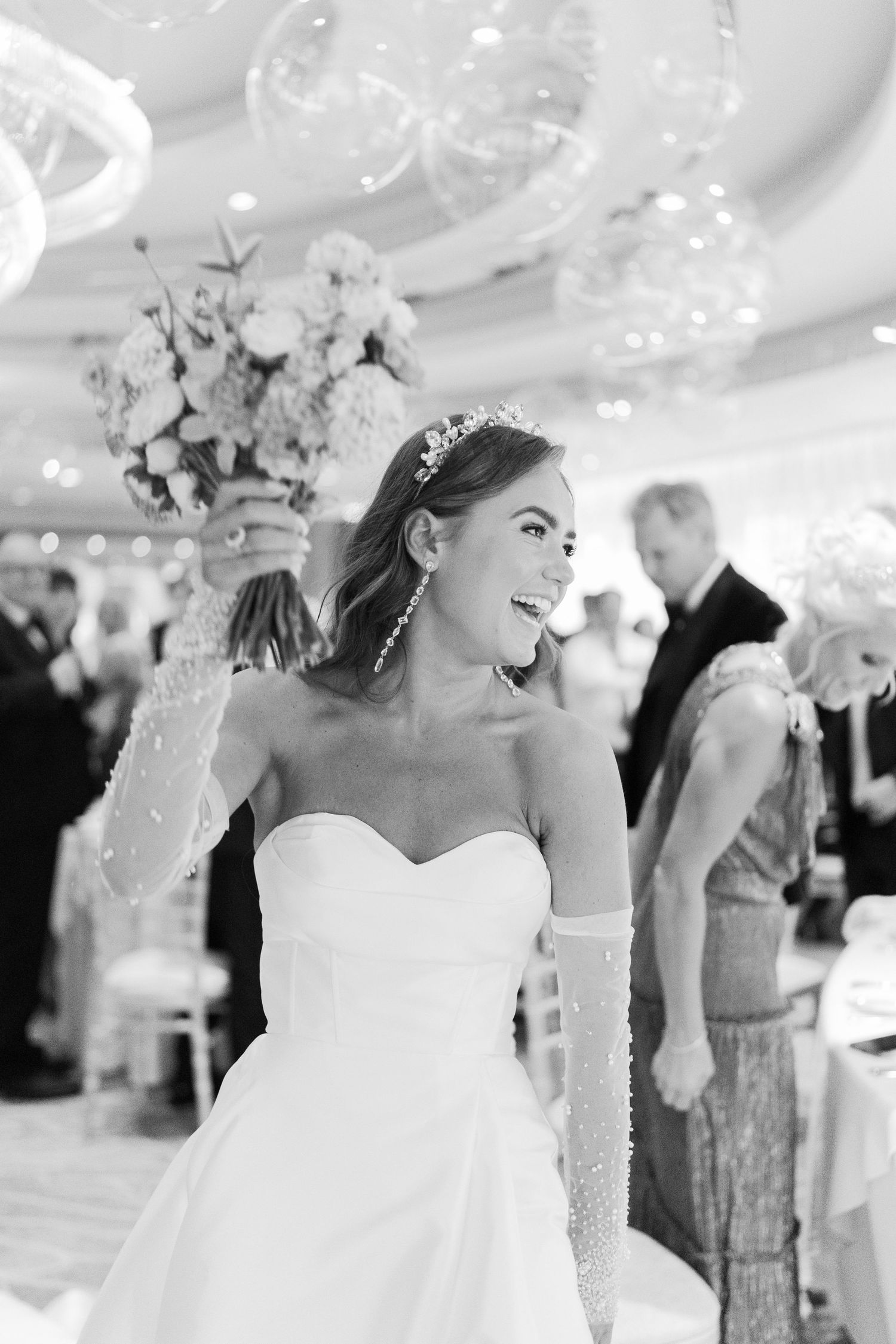 Black and white photo of a smiling bride in a sweetheart gown with pearl gloves and tiara at an elegant wedding reception.