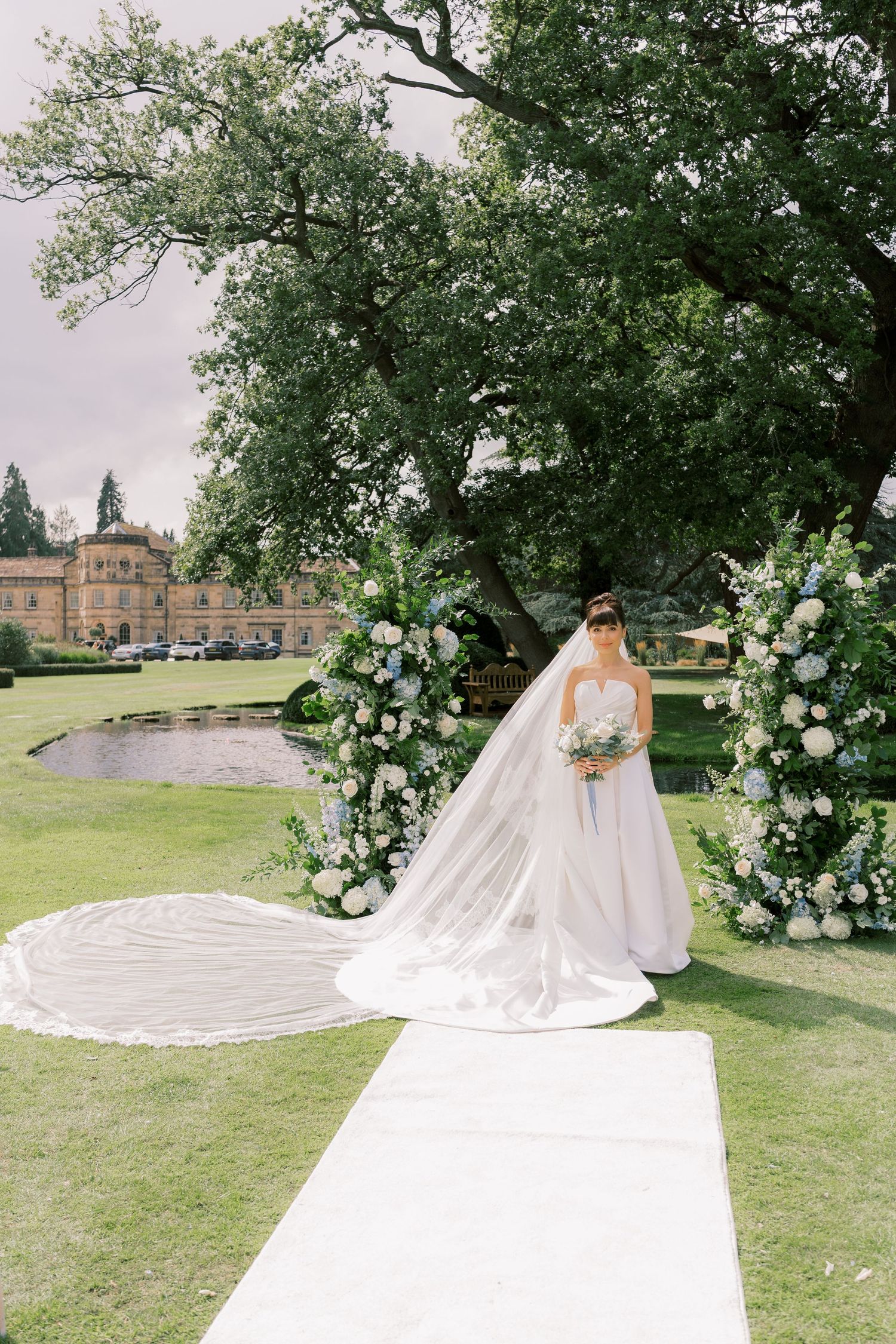 Bride in a white ballgown with long veil stands at floral arch altar on green lawn with historic manor backdrop.