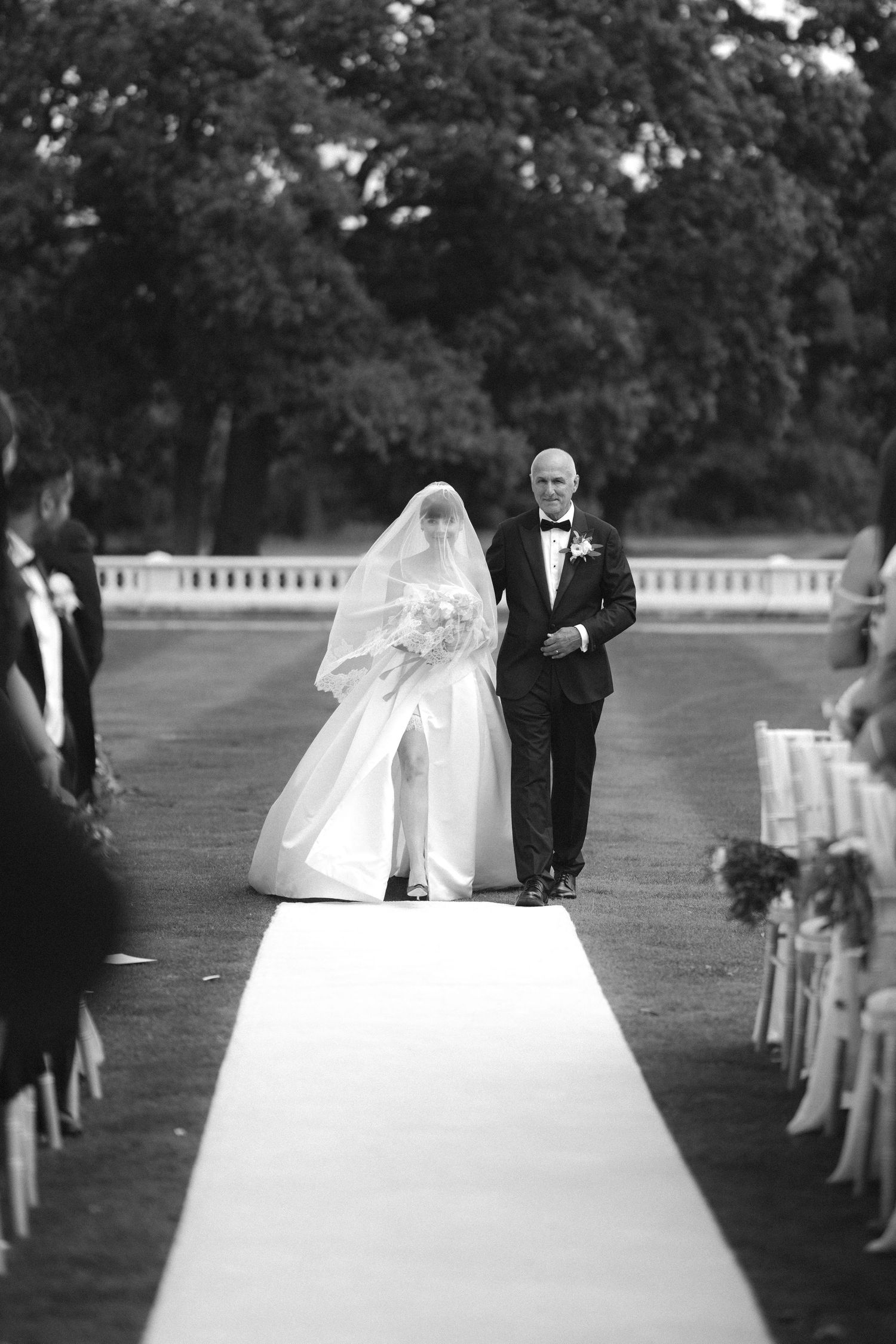 Black and white photo of a bride walking down the aisle with an older man in a tuxedo at an outdoor wedding ceremony.