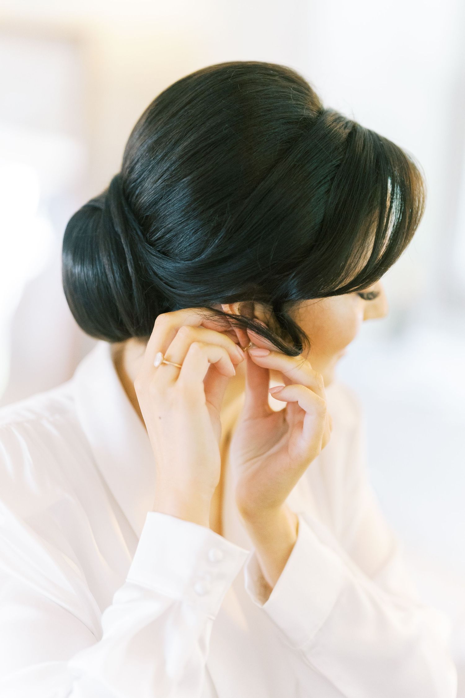 Bride in white shirt adjusting earring with elegant dark updo hairstyle during wedding preparations.