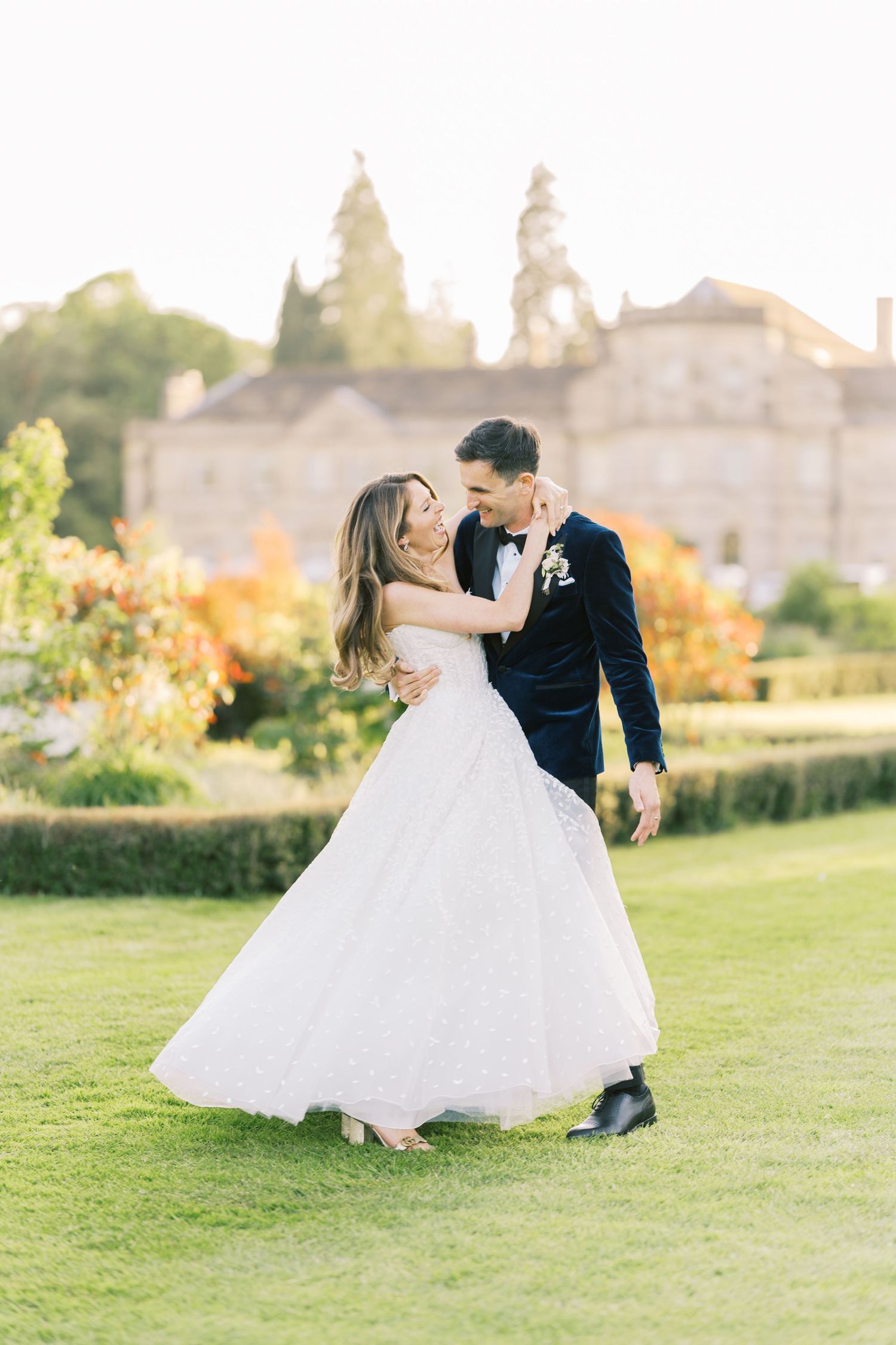 Bride and groom dance joyfully on green lawn with colorful autumn gardens and historic manor in background.