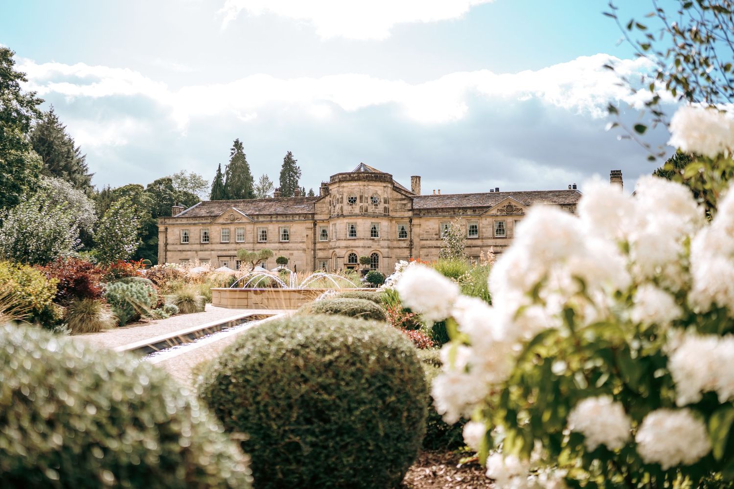 Elegant Italian villa surrounded by manicured gardens with white roses and sculpted hedges under a partly cloudy sky.