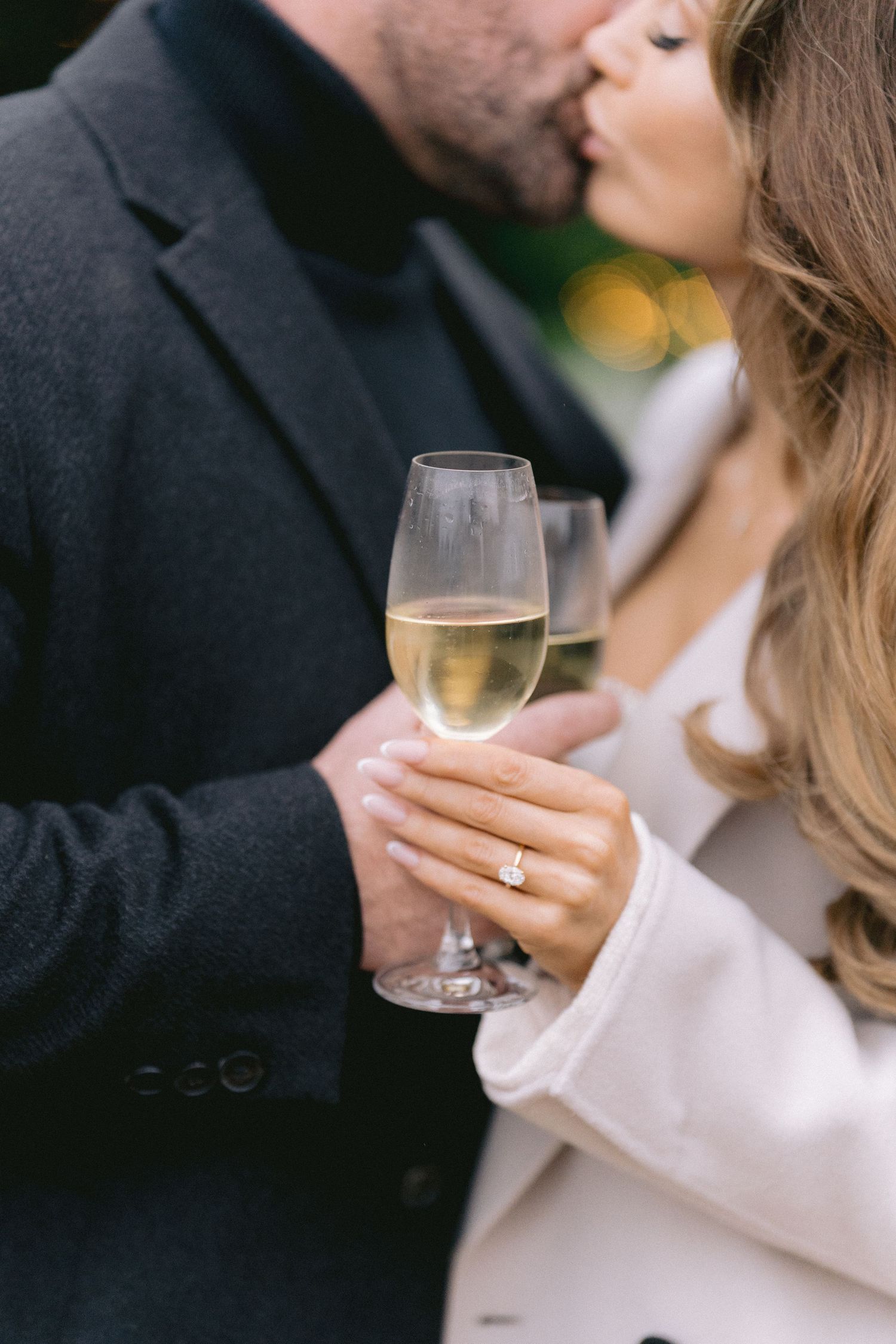 Newly engaged couple kisses while holding champagne glasses, diamond ring visible on womans finger.