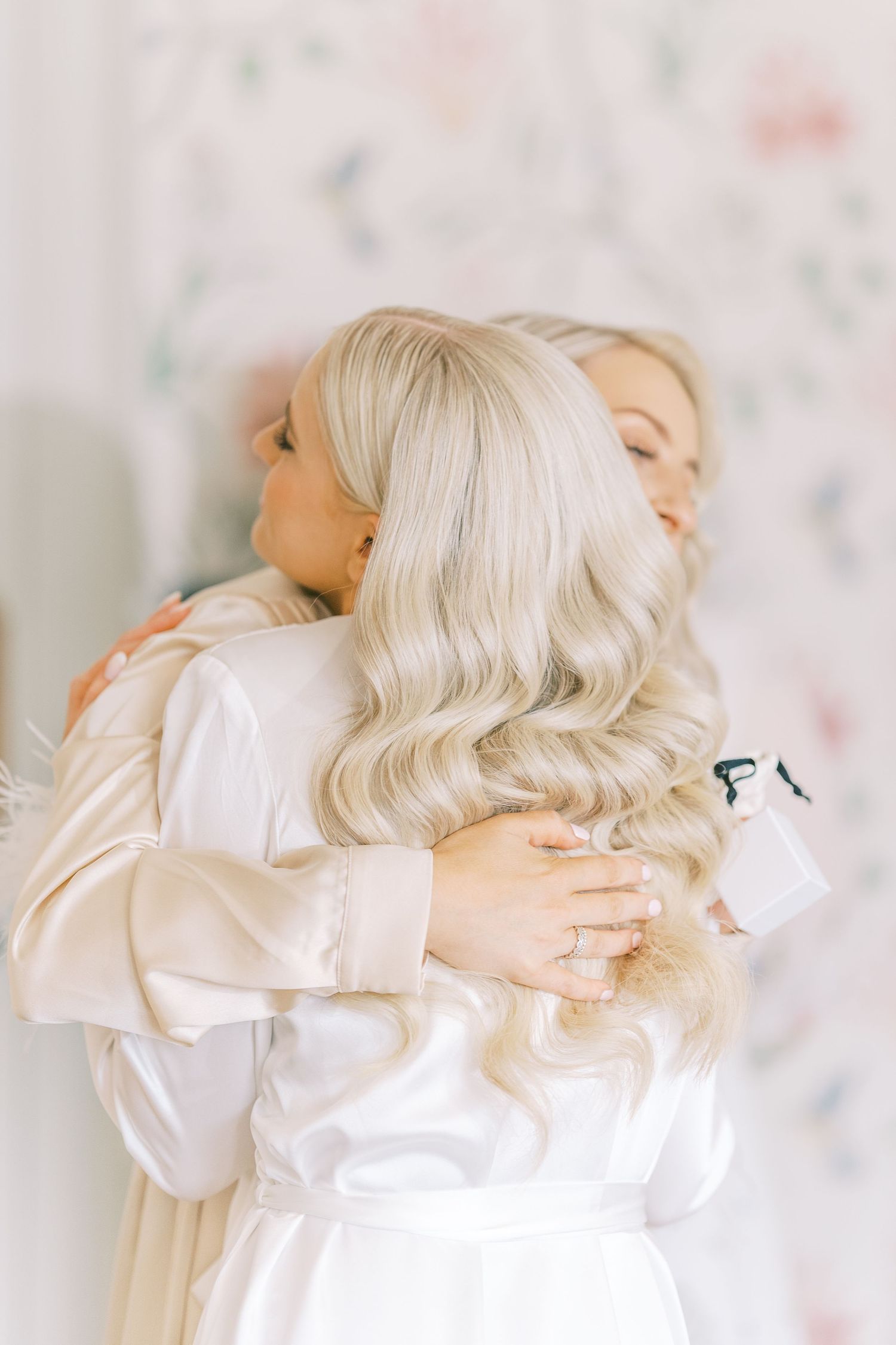Two blonde women in silk robes sharing an emotional embrace during bridal preparations.