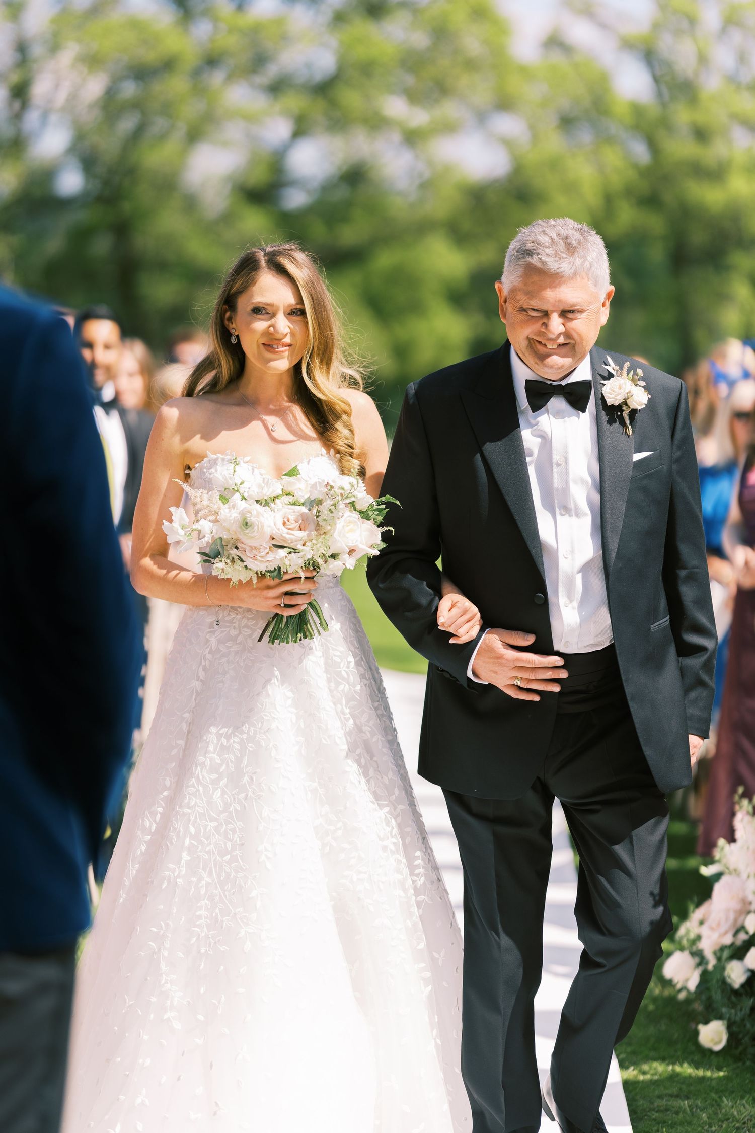 Bride and father walking down outdoor wedding aisle in sunshine.